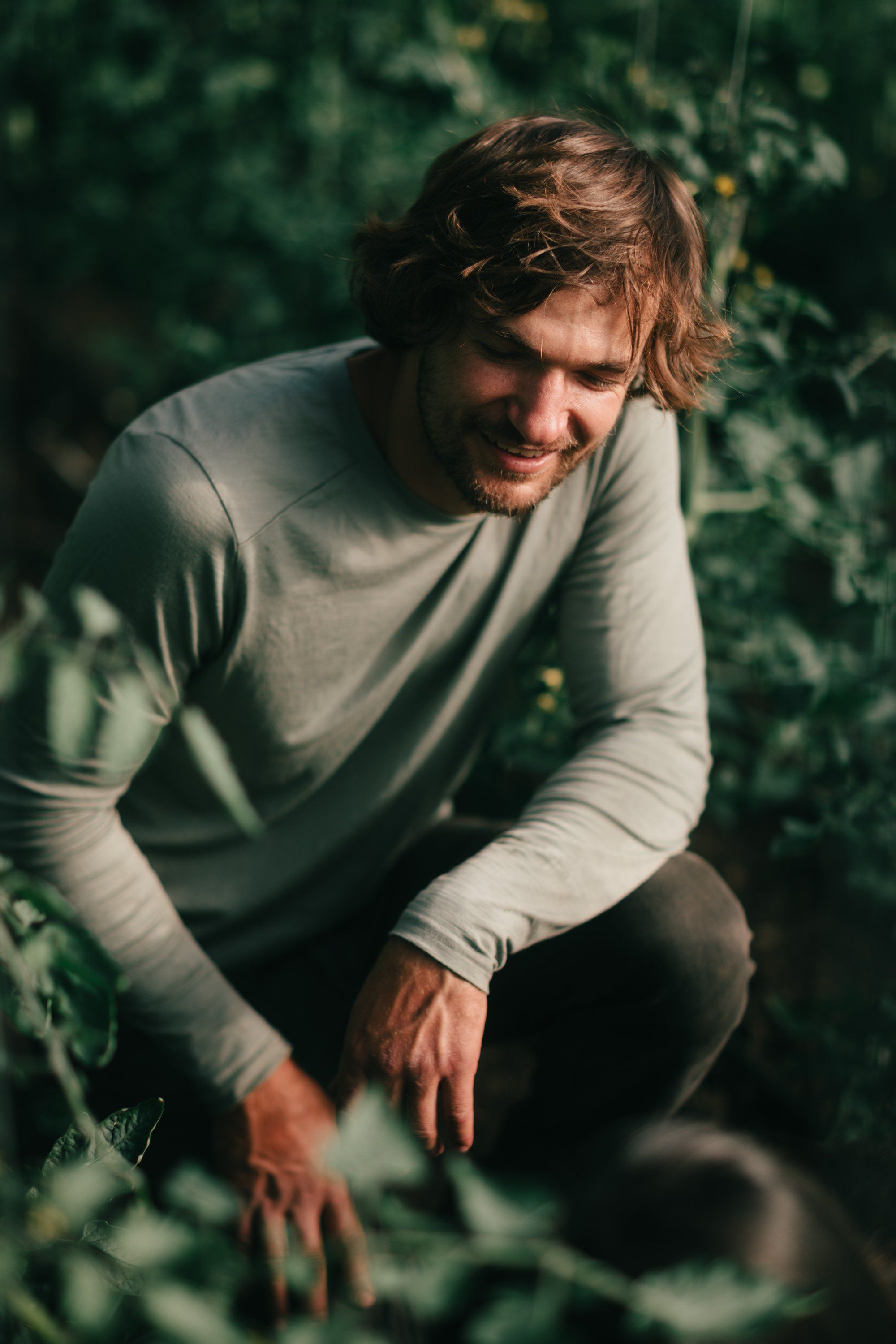 Man in cream long-sleeve shirt crouching among lush green foliage, warm sunlight filtering through leaves