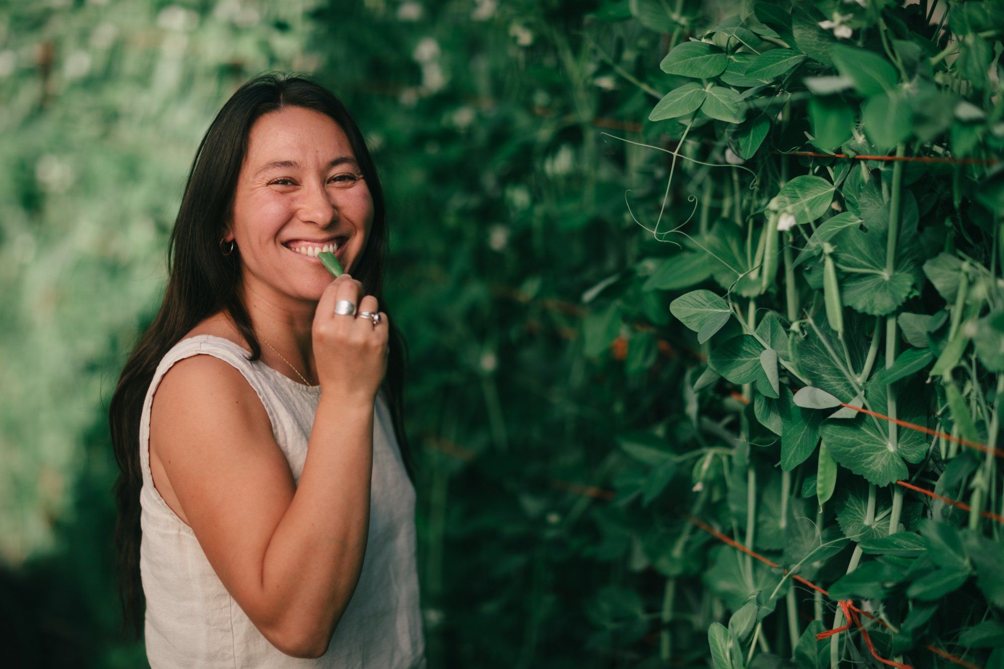 Woman in white sleeveless top smiling beside lush green garden vines in natural daylight