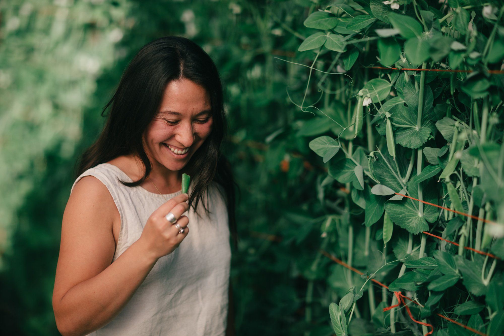 Woman in light gray tank top smiling while standing among lush green garden vines and foliage