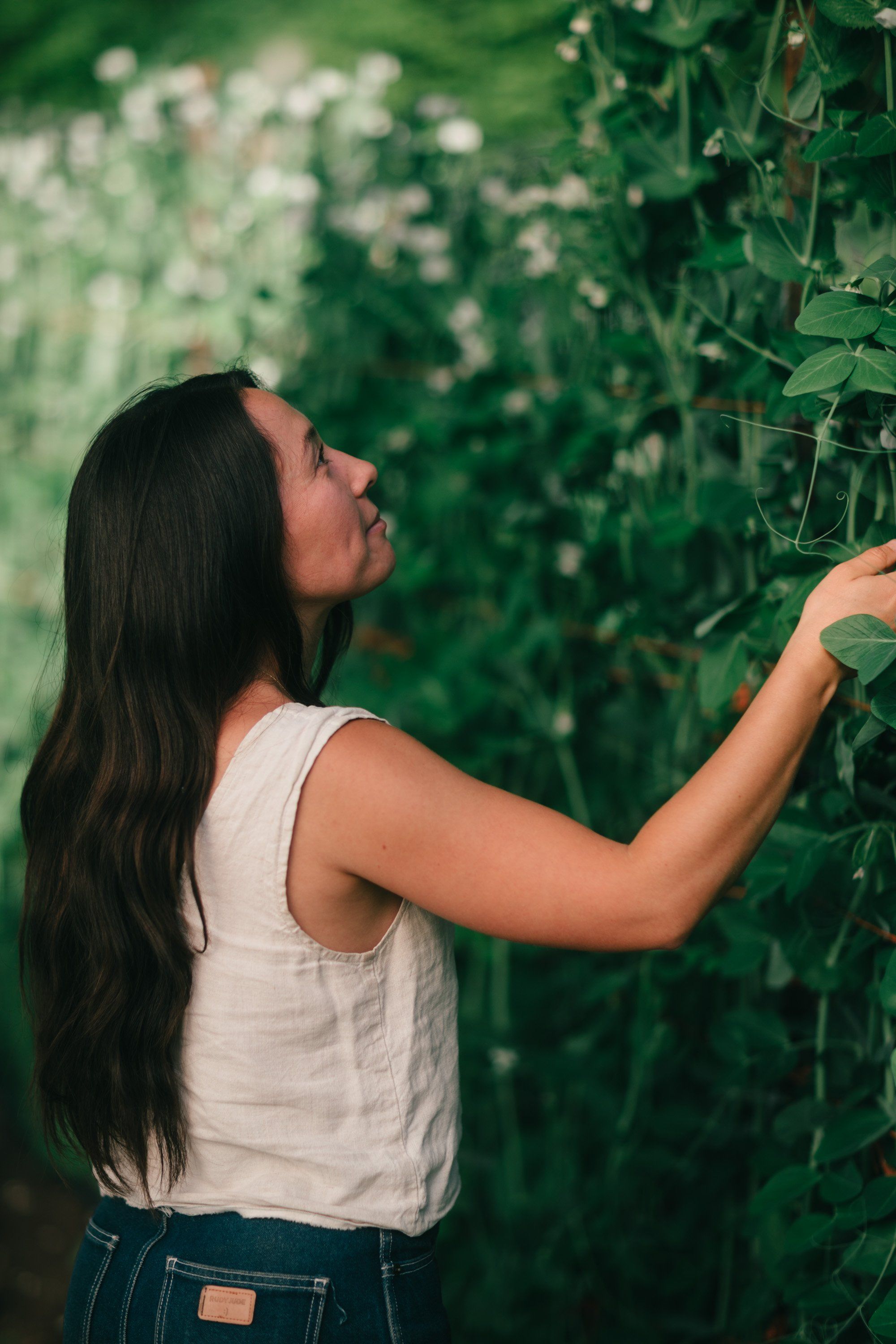 Woman in white tank top reaching toward leafy green vines in golden afternoon light on a farm