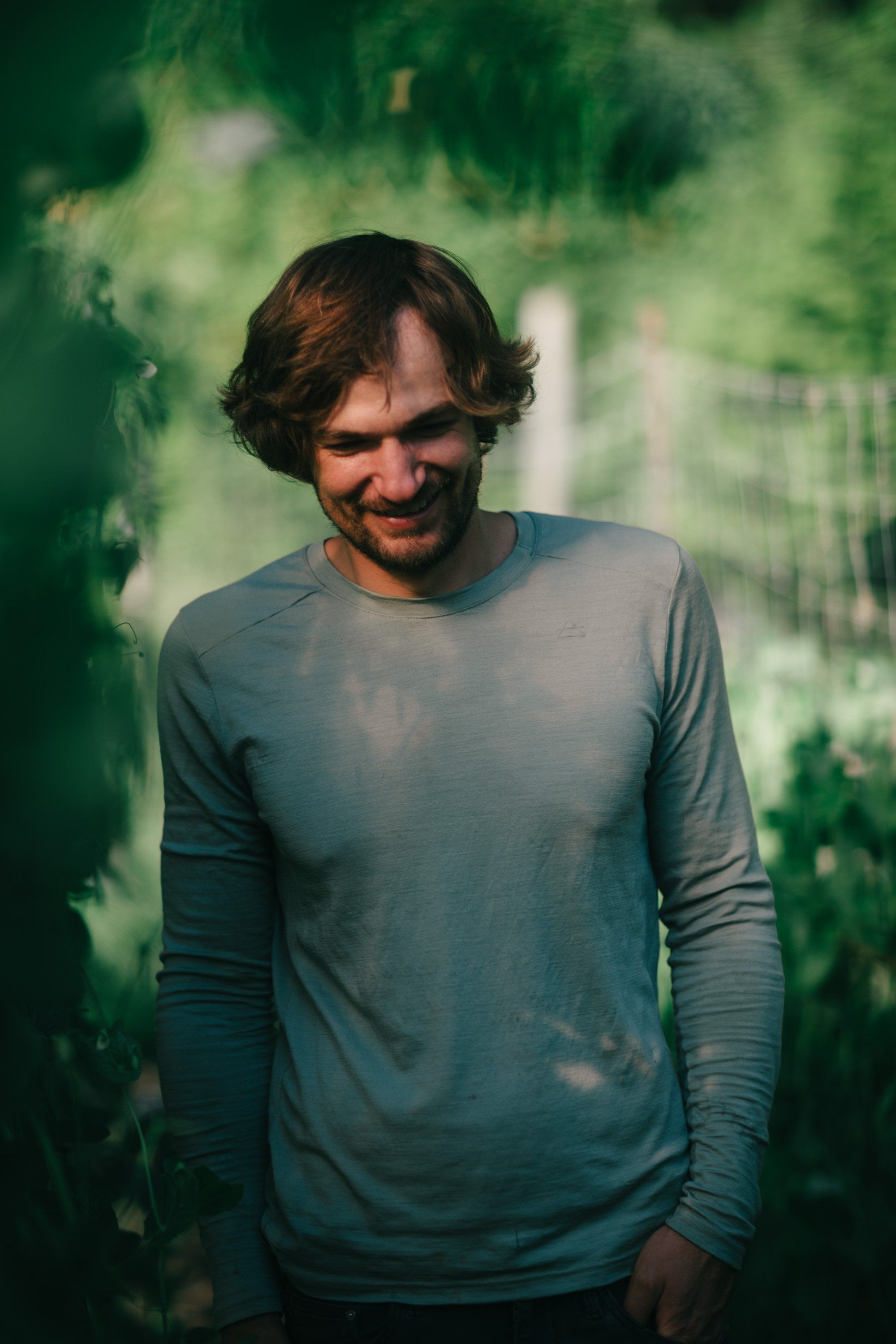 Man with shoulder-length dark hair smiling against lush green foliage, wearing a dusty blue long-sleeve shirt
