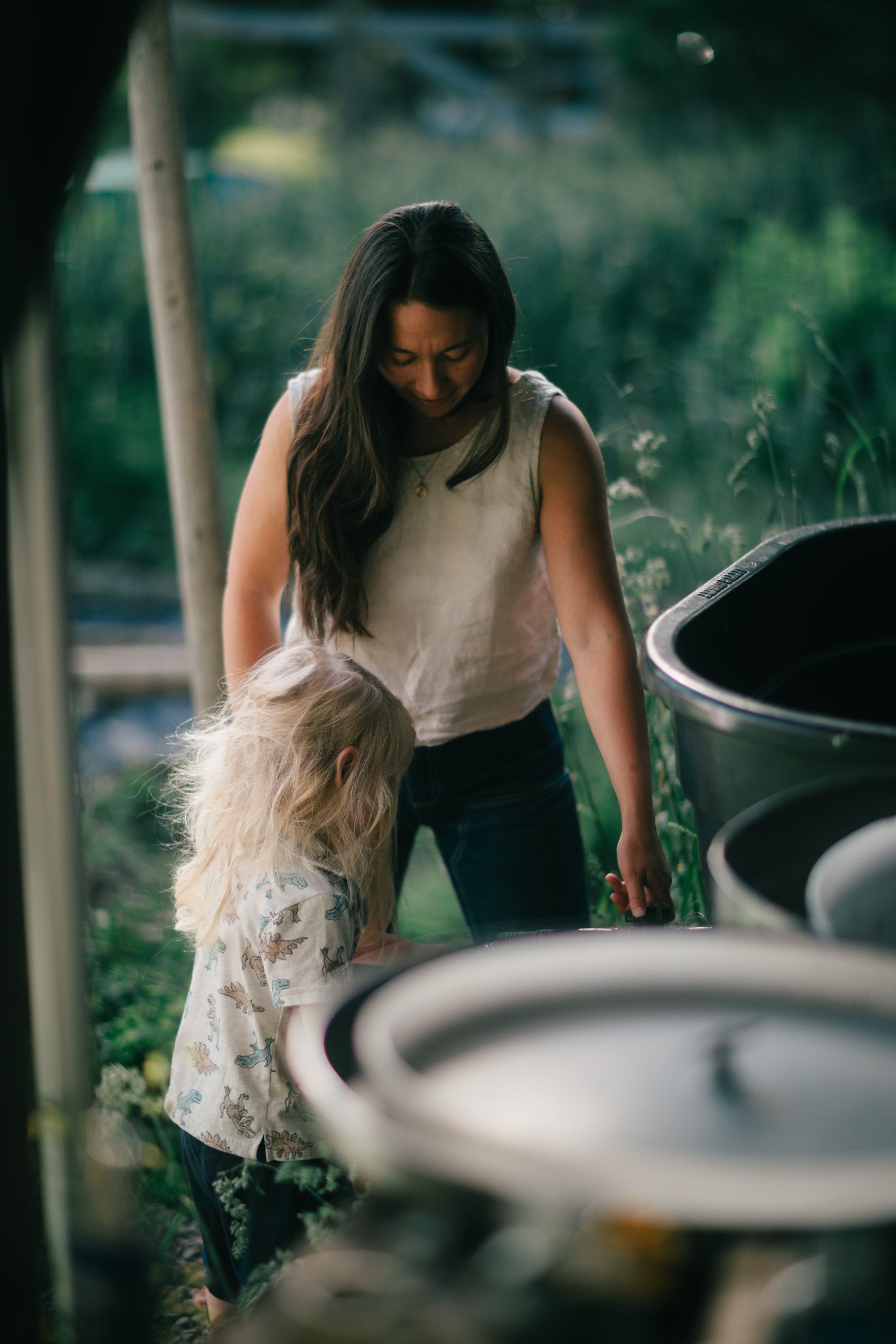 Woman in cream sleeveless top bending toward small child in lace dress among garden planters, dappled woodland light