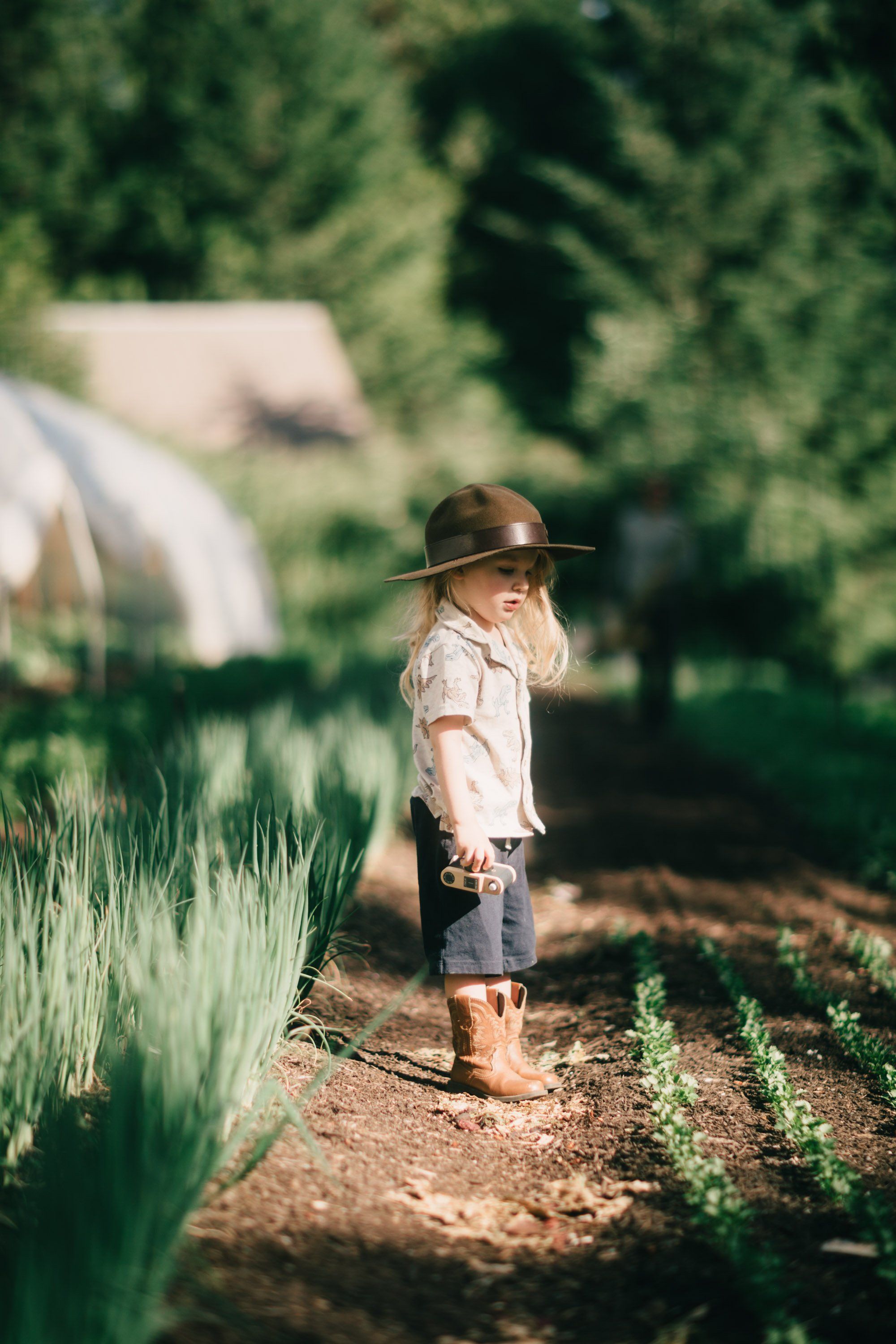 Child in cowboy hat walking barefoot down garden path lined with green plants and raised beds
