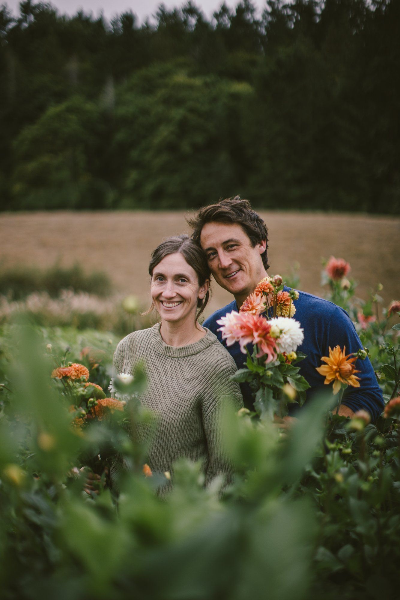 Couple smiling among blooming dahlias in a flower field, evergreens behind, golden afternoon light