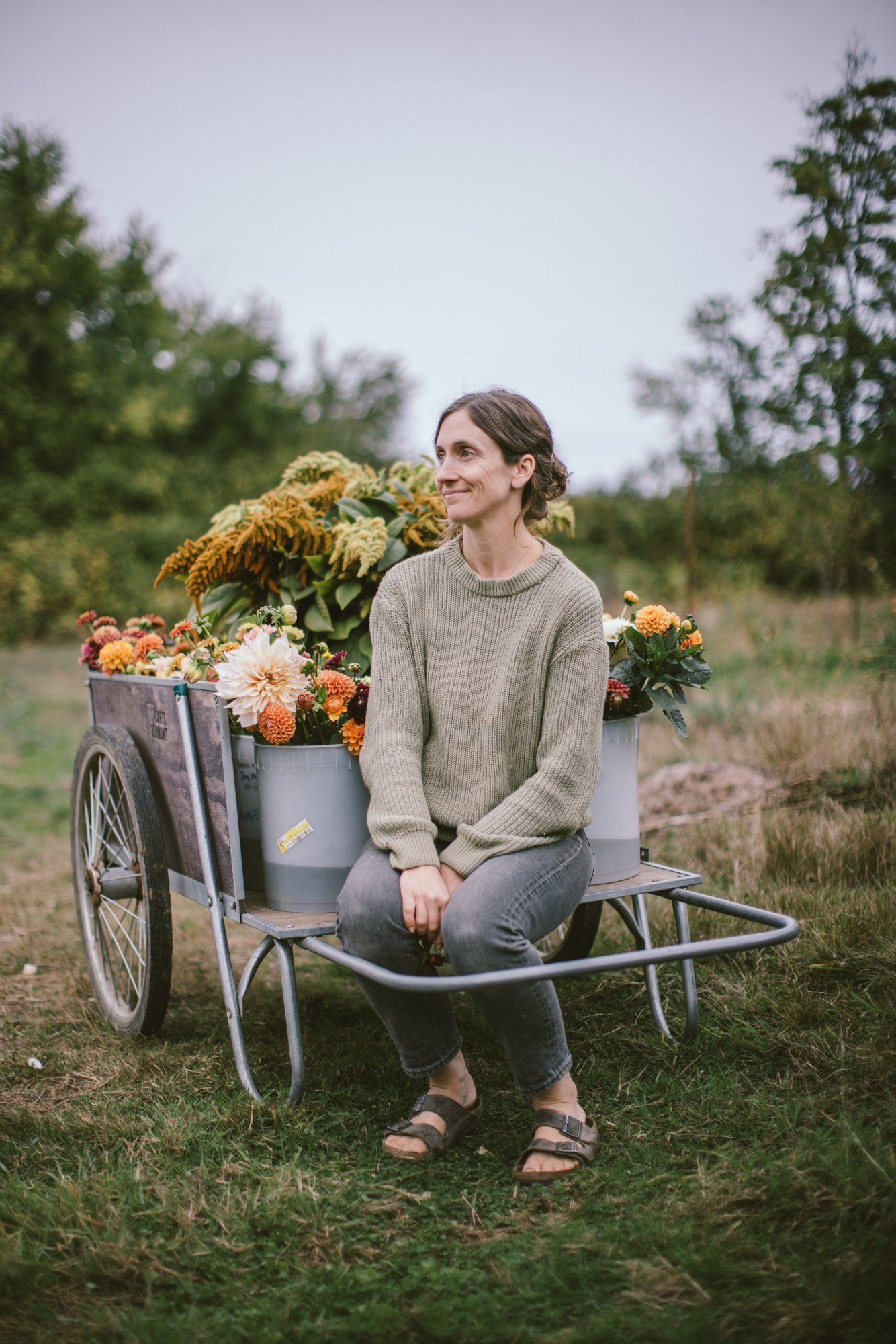 Woman in beige sweater sits on farm cart filled with golden and orange flowers at Bullock Lake Farm