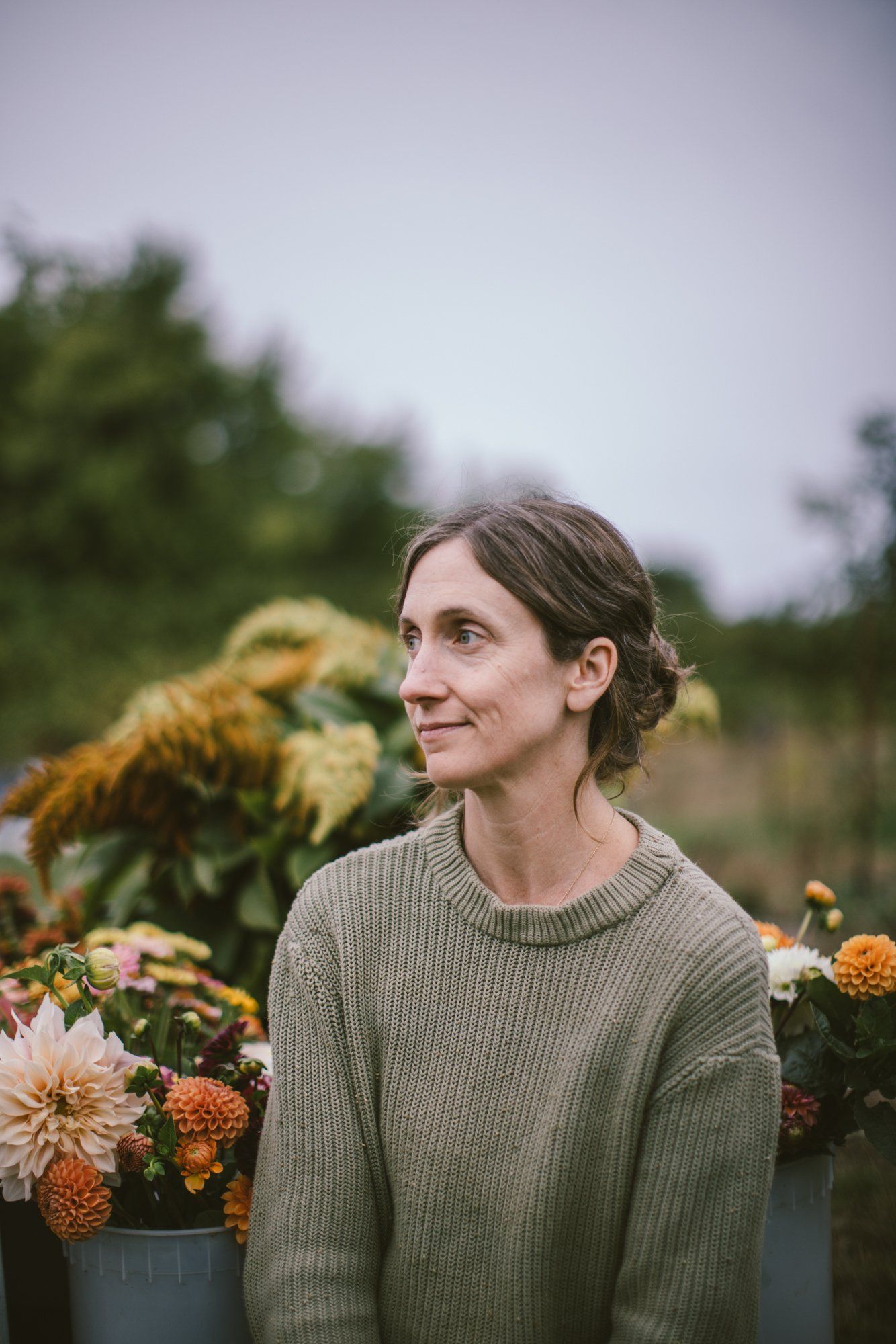 Woman in sage green sweater gazes thoughtfully beside blooming dahlias at a farm garden
