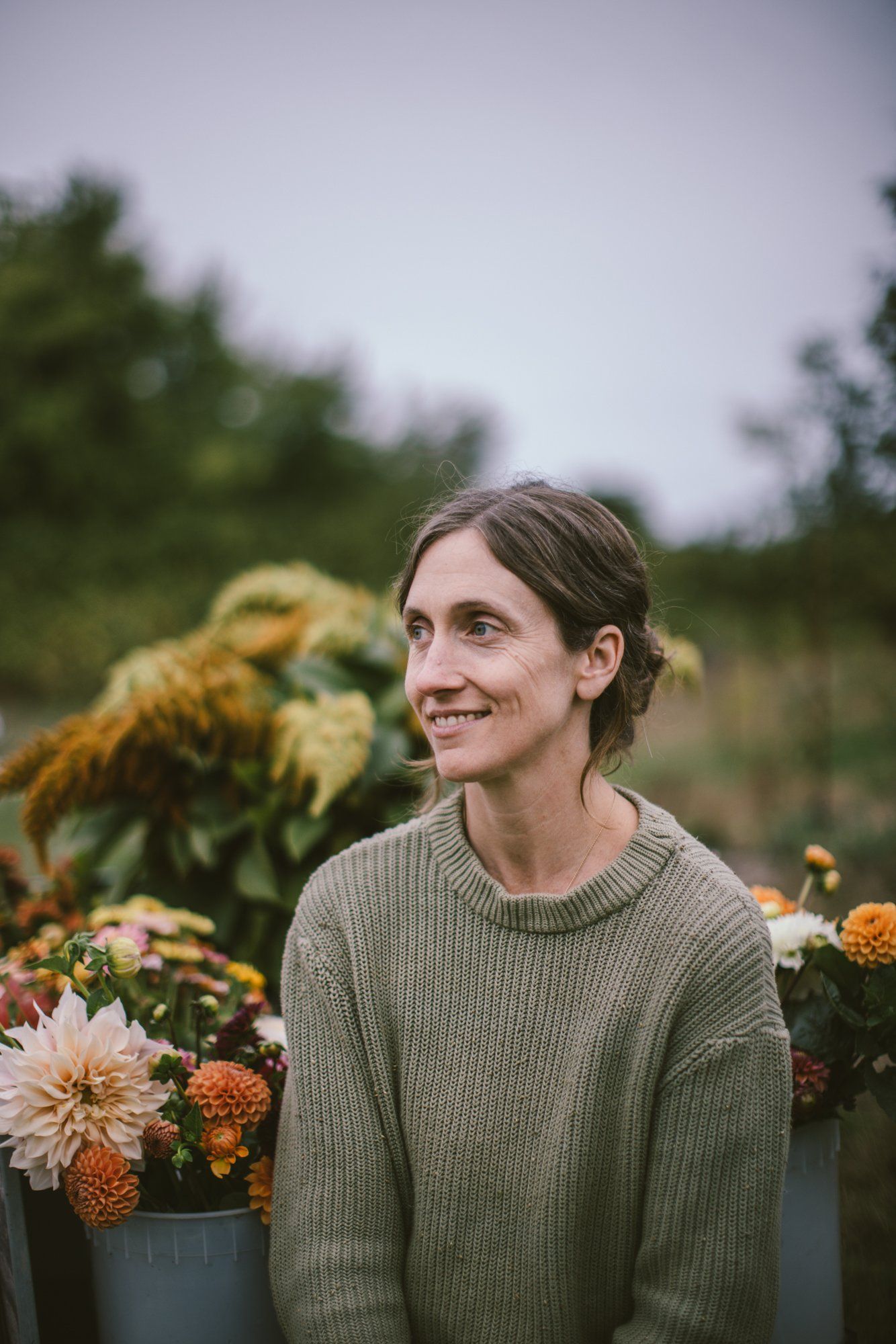 Woman in sage green ribbed sweater smiling beside orange and cream dahlias at Bullock Lake Farm