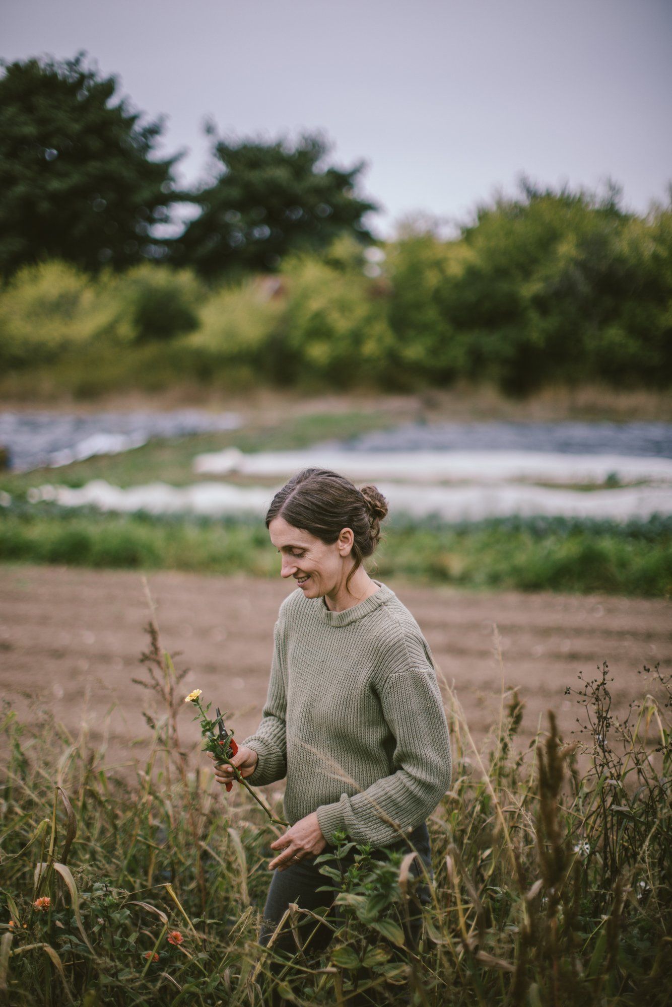 Woman in cream sweater standing among dried grasses with river and tree-lined banks behind her on overcast day