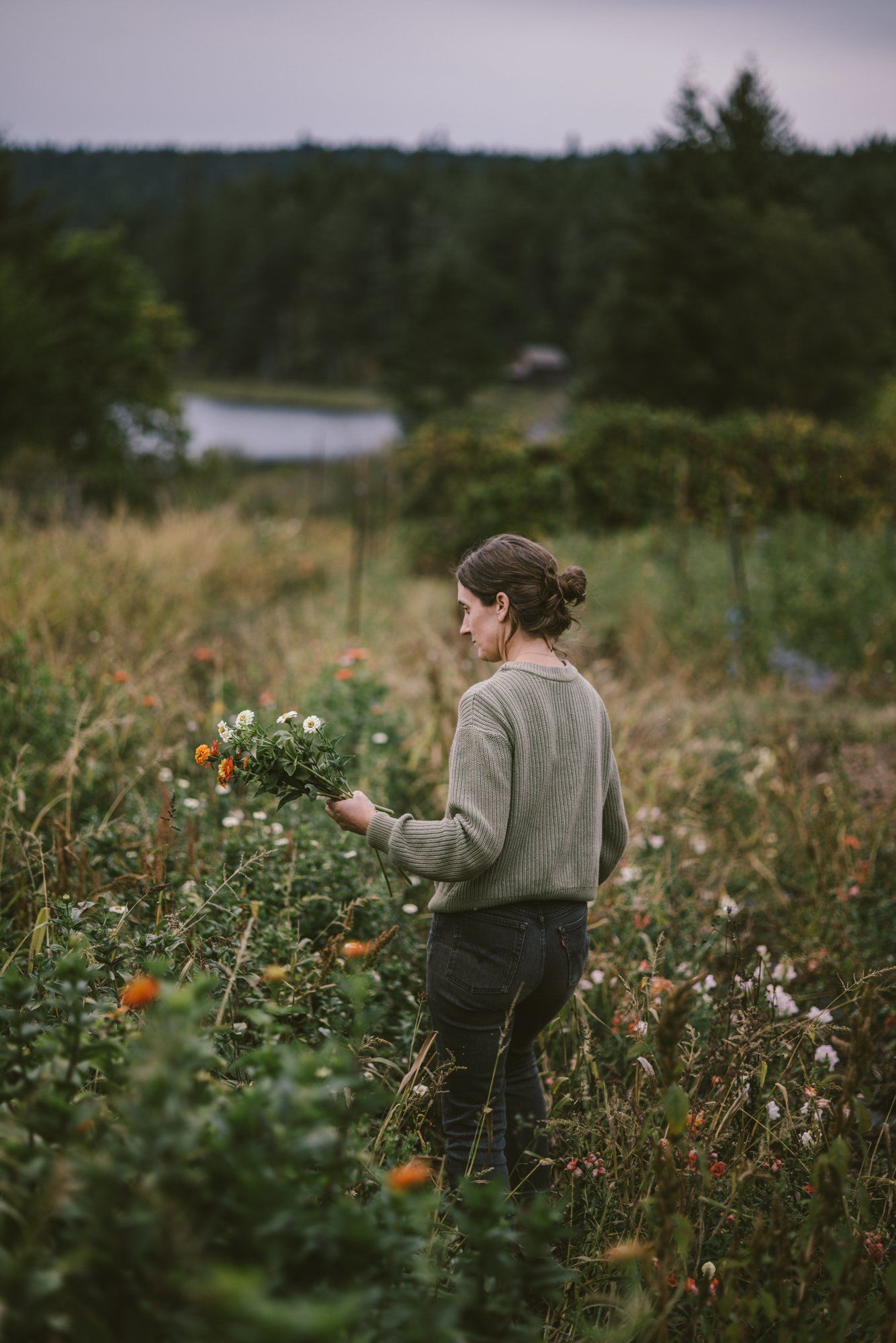 Woman in cream cardigan picking wildflowers in a meadow with forest and pond beyond at dusk