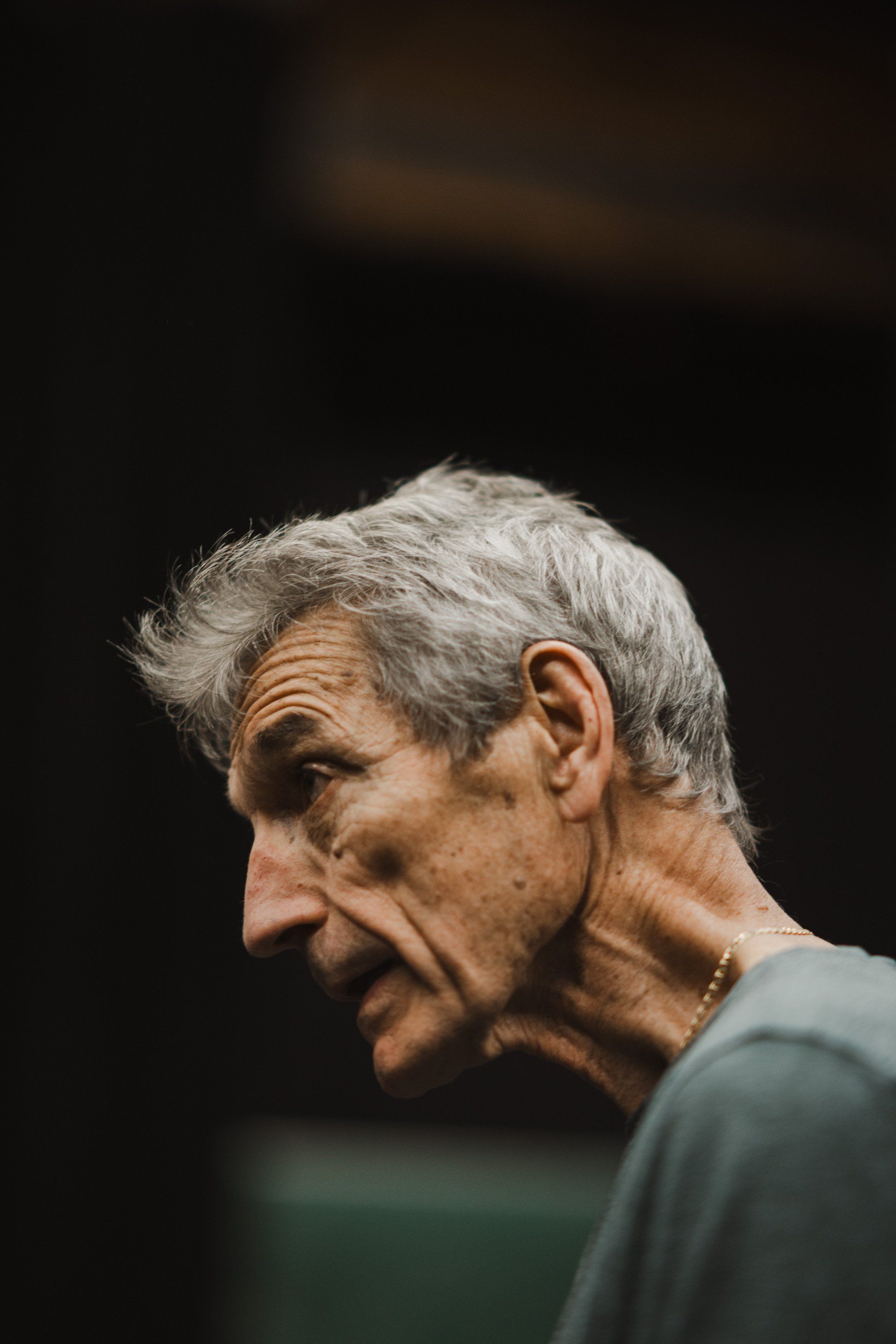 Elderly man in profile against dark background, gray hair catching warm light, gentle expression, wearing gray shirt
