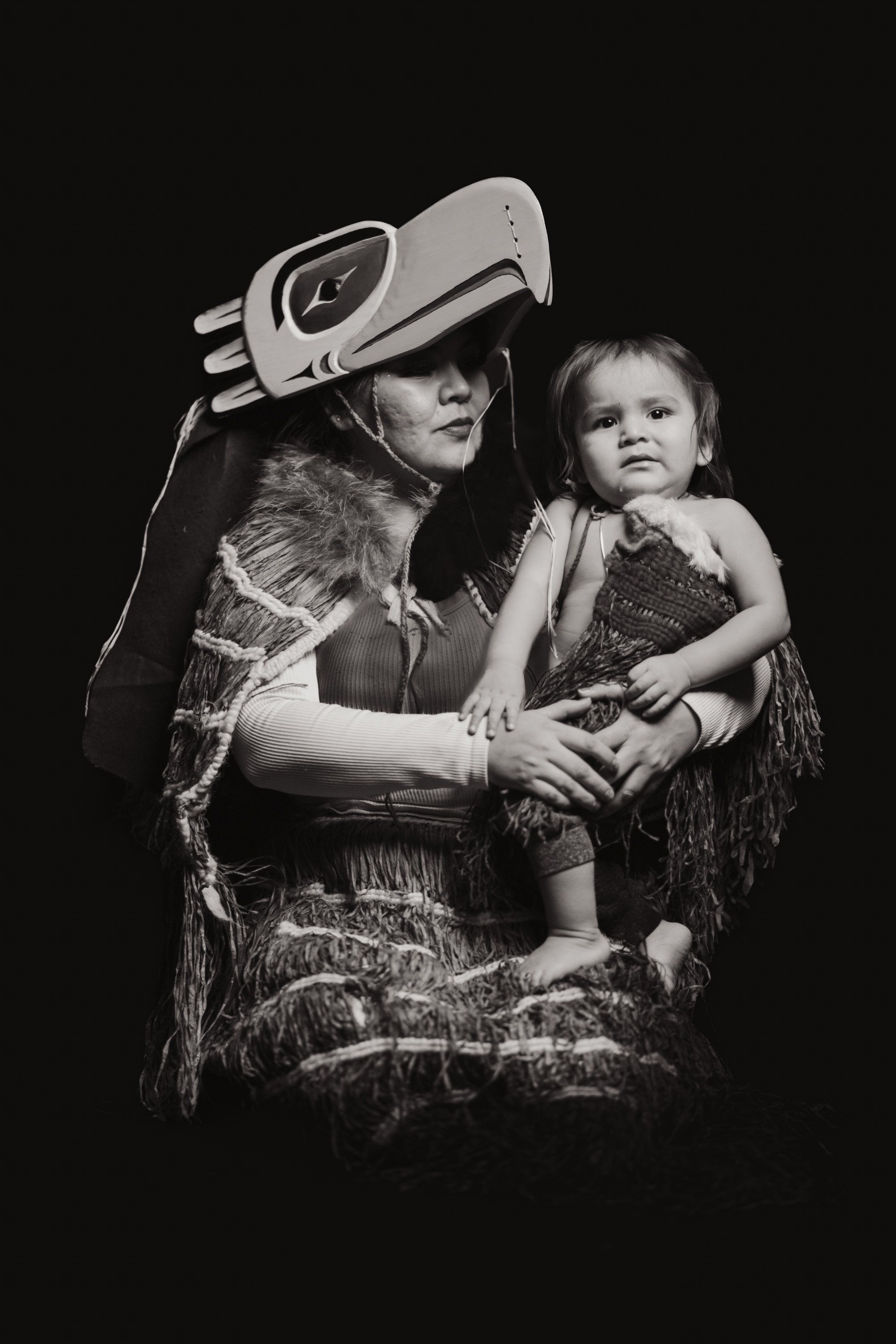 Woman wearing traditional regalia and carved hat holds a young child against dark background, their faces close in an intimate portrait