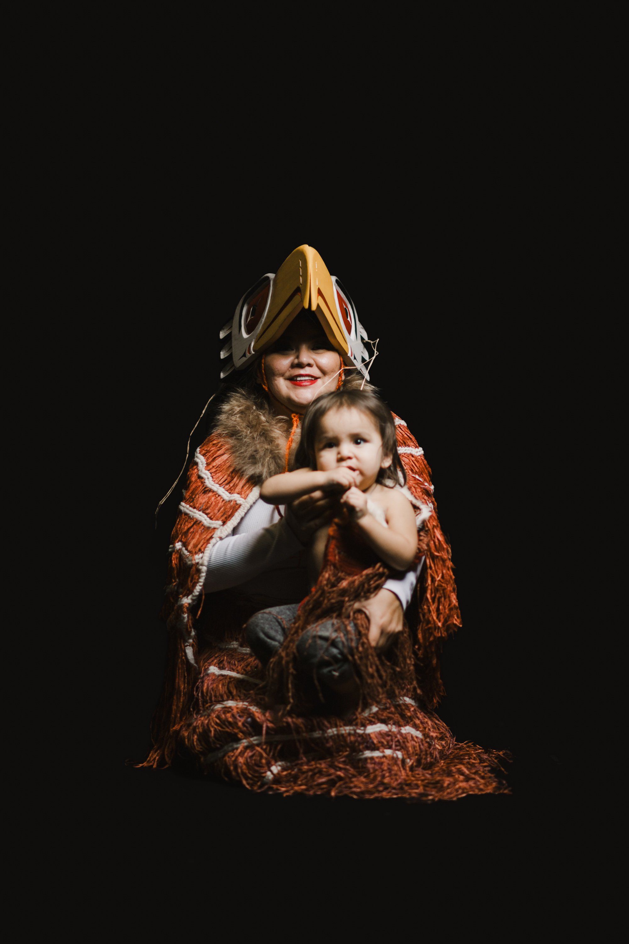 Adult and child in traditional regalia with fur and cedar, posed against black background with dramatic side lighting