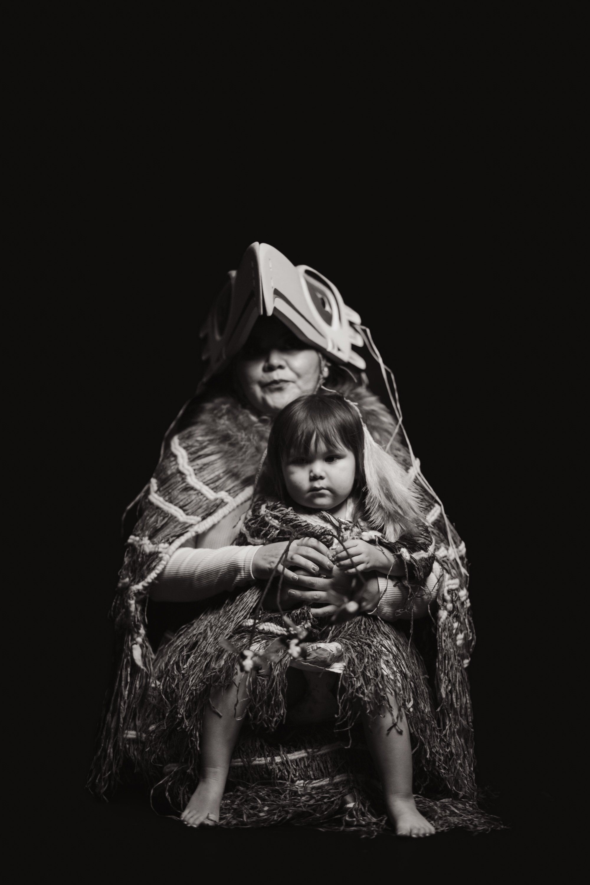 Adult and child wrapped together in patterned blanket against black background, intimate portrait celebrating Coast Salish family connection