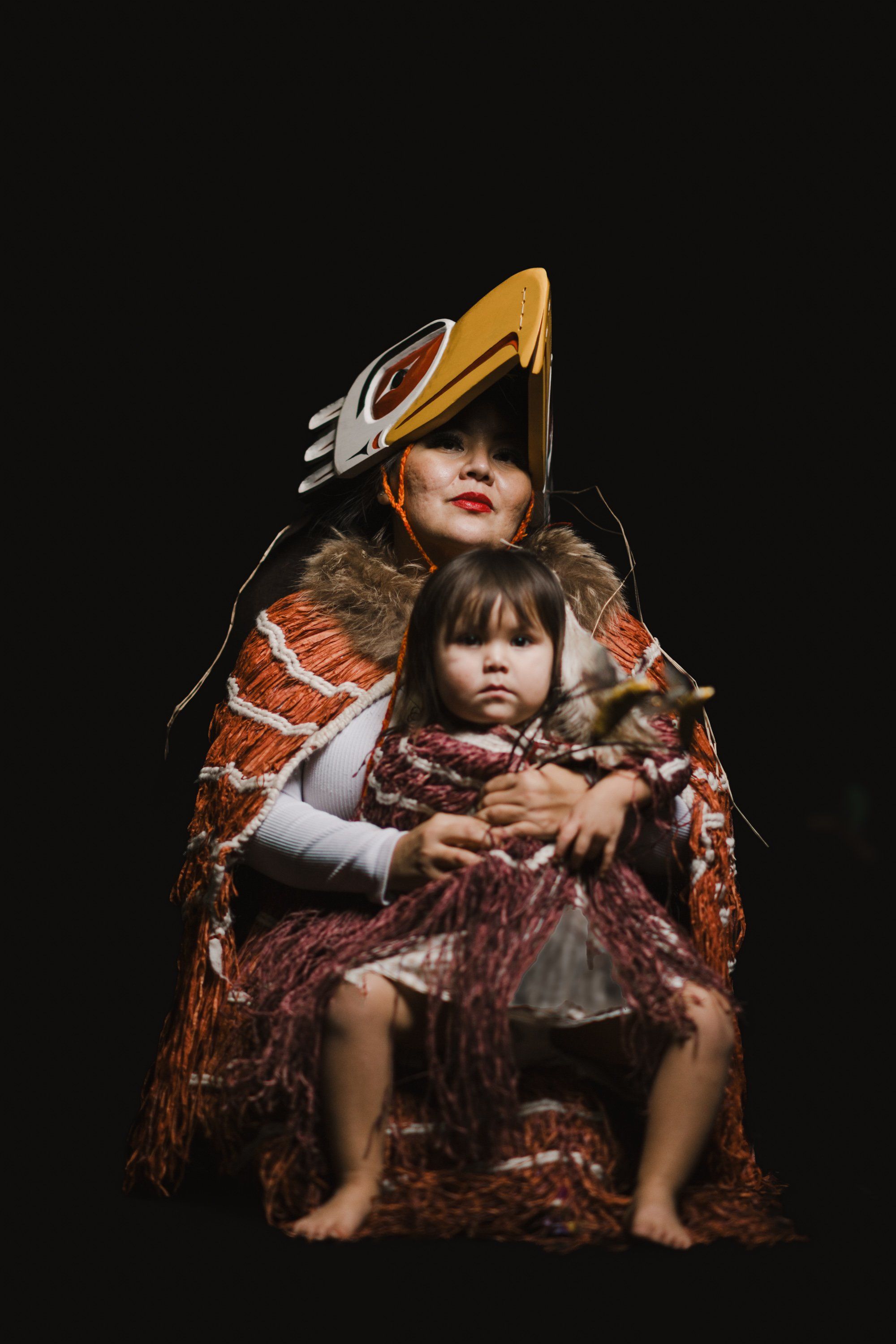 Adult and child in traditional regalia with copper and red woven blankets against black background, wearing distinctive gold ceremonial hat