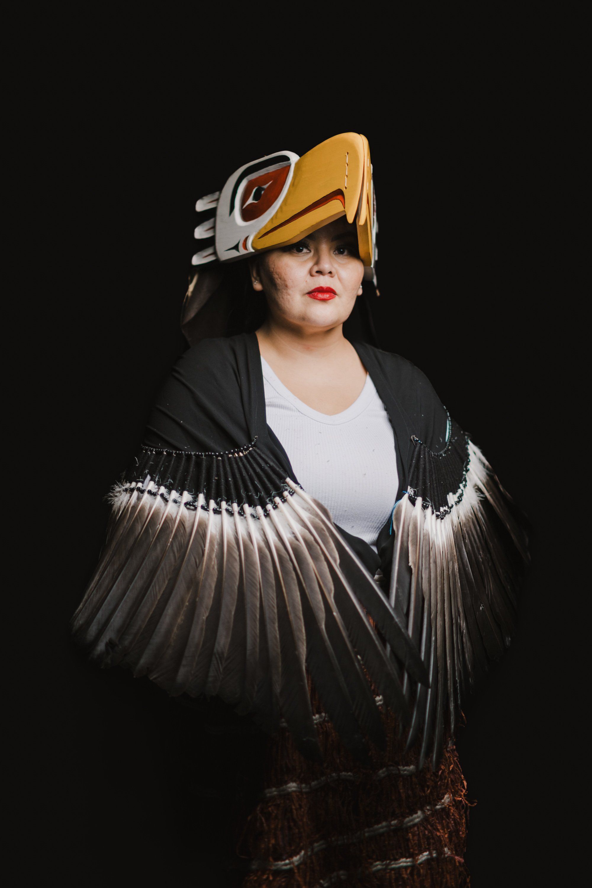 Person wearing gold ceremonial hat and dramatic black feathered wings over dark jacket against black background, striking theatrical pose