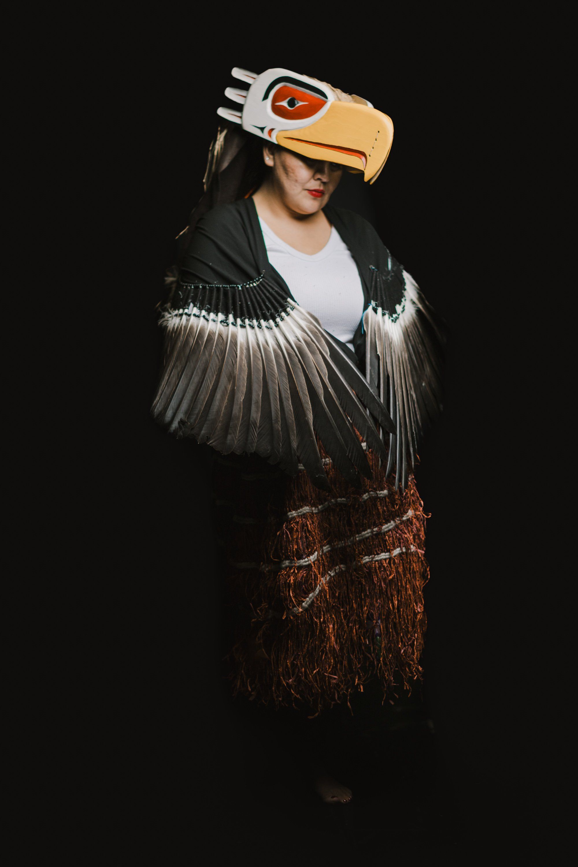 Person wearing traditional Coast Salish regalia with carved eagle mask, silver-lined wings, and woven skirt against black background