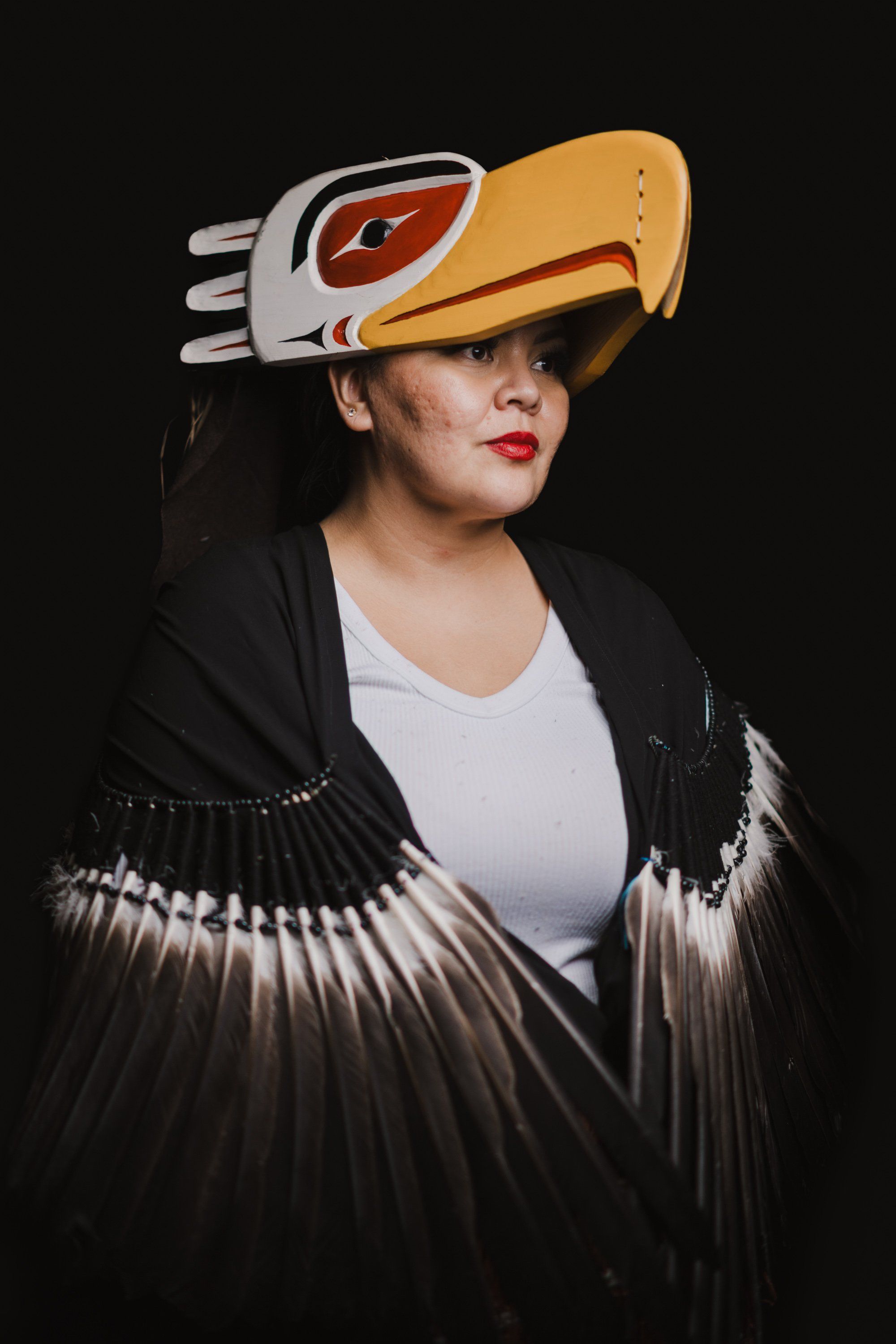 Person wearing dramatic Coast Salish regalia with carved eagle headdress and fringed black button blanket against dark background