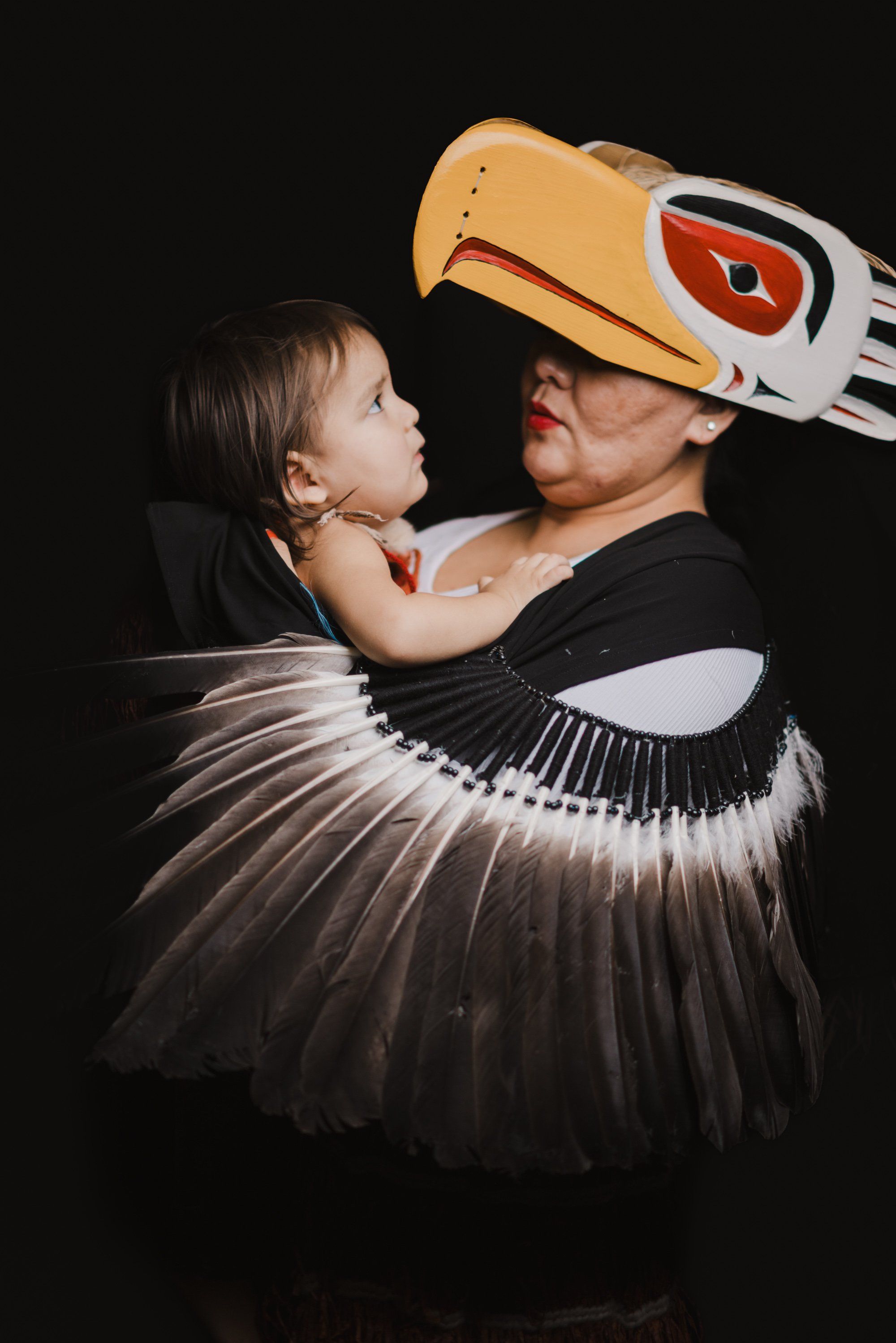 Adult in traditional regalia with carved eagle headdress holds child against dramatic black background, their faces close in intimate moment