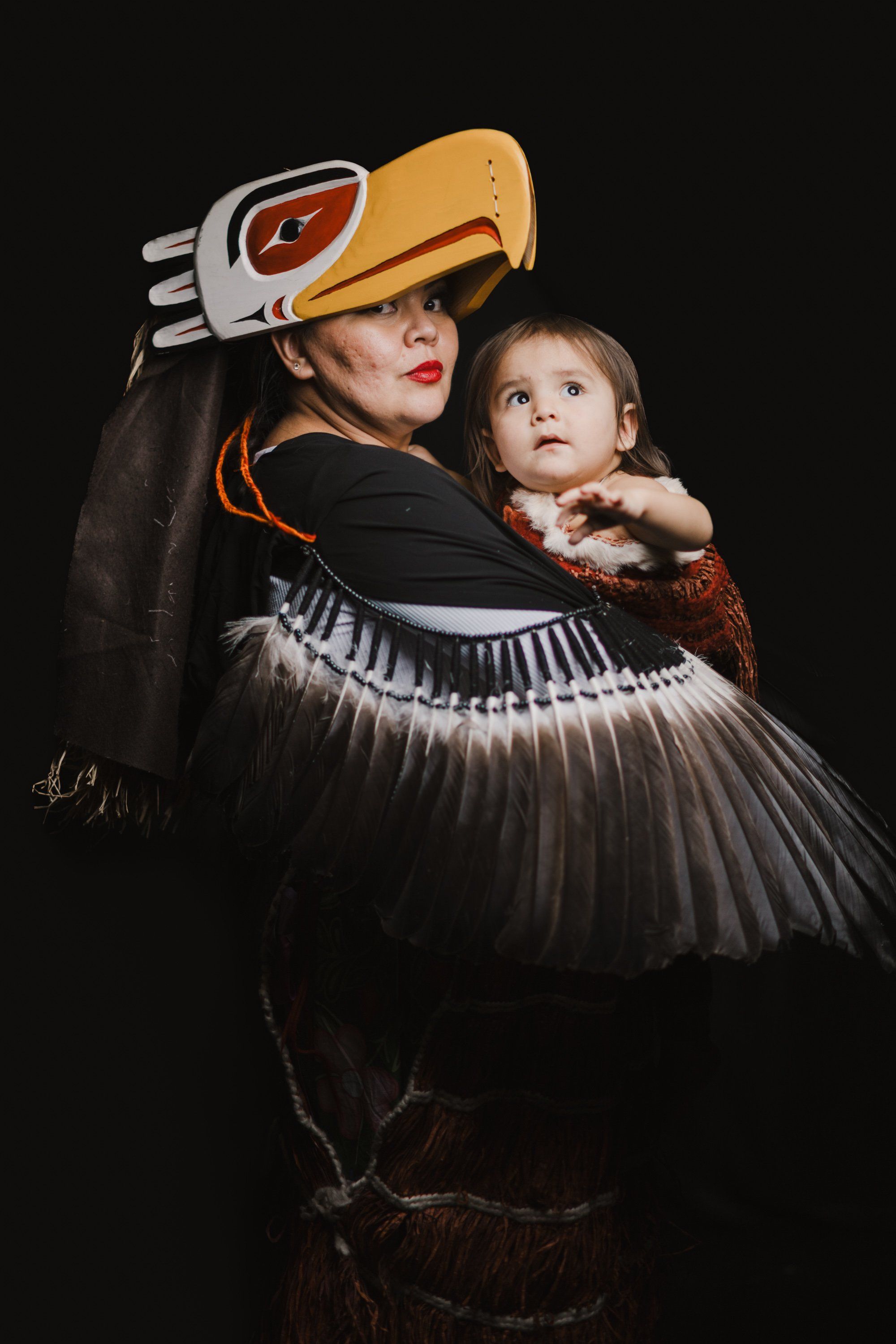 Adult in orange and white regalia holding child against dark background, both gazing upward in contemplative pose