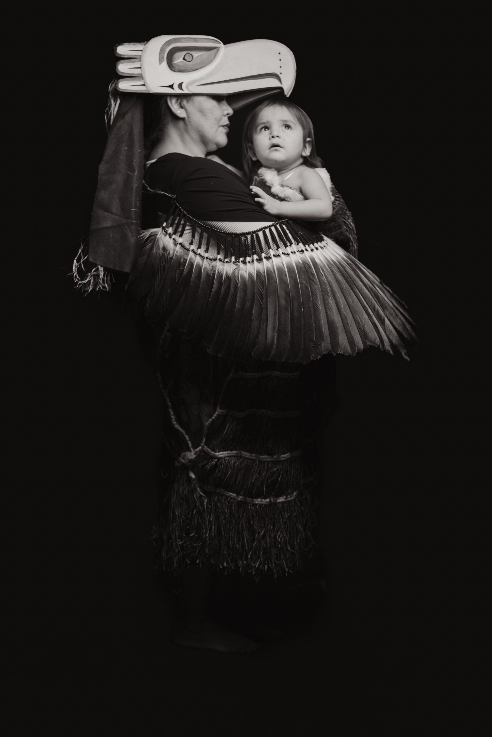 Woman in traditional Coast Salish regalia holding a child against a black background, dramatic side lighting