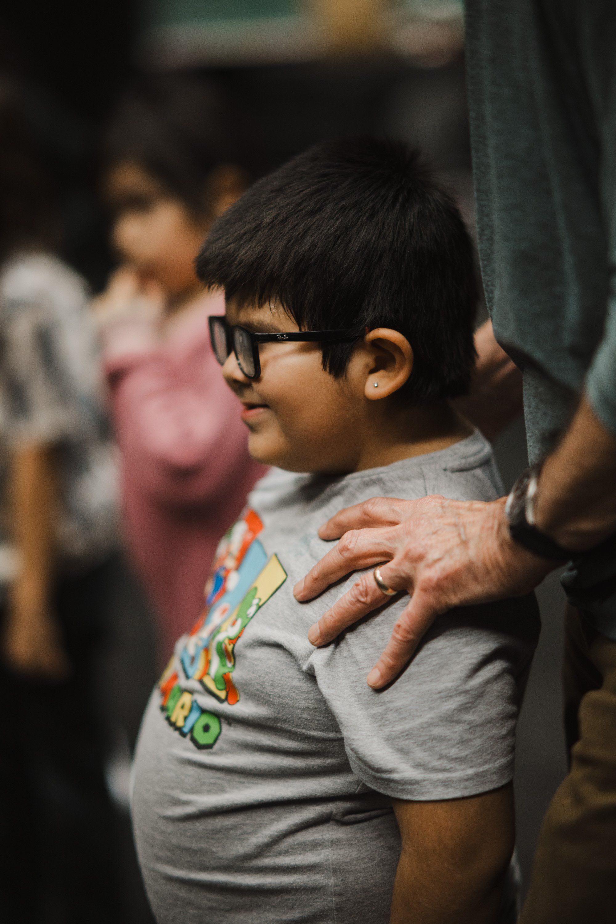 Young boy in glasses wearing a gray graphic tee stands close beside an adult, smiling softly in warm indoor light