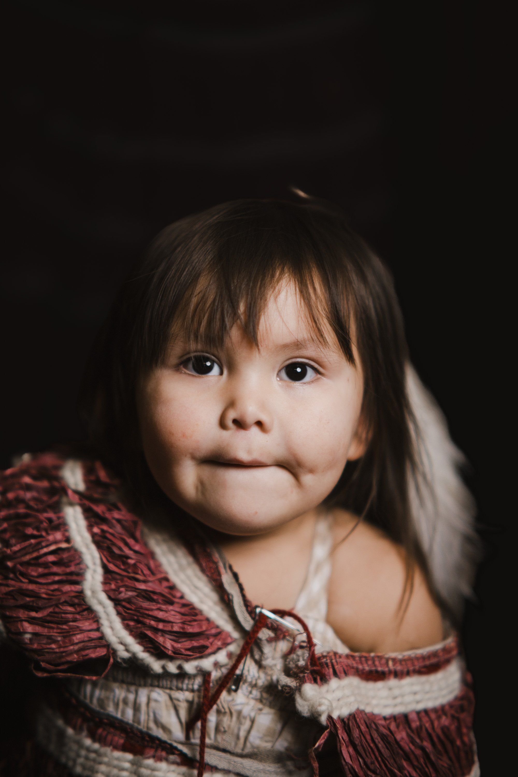 Young child with dark hair and bangs wearing a burgundy and cream patterned garment against a dark background, gazing directly at camera with a serene expres…