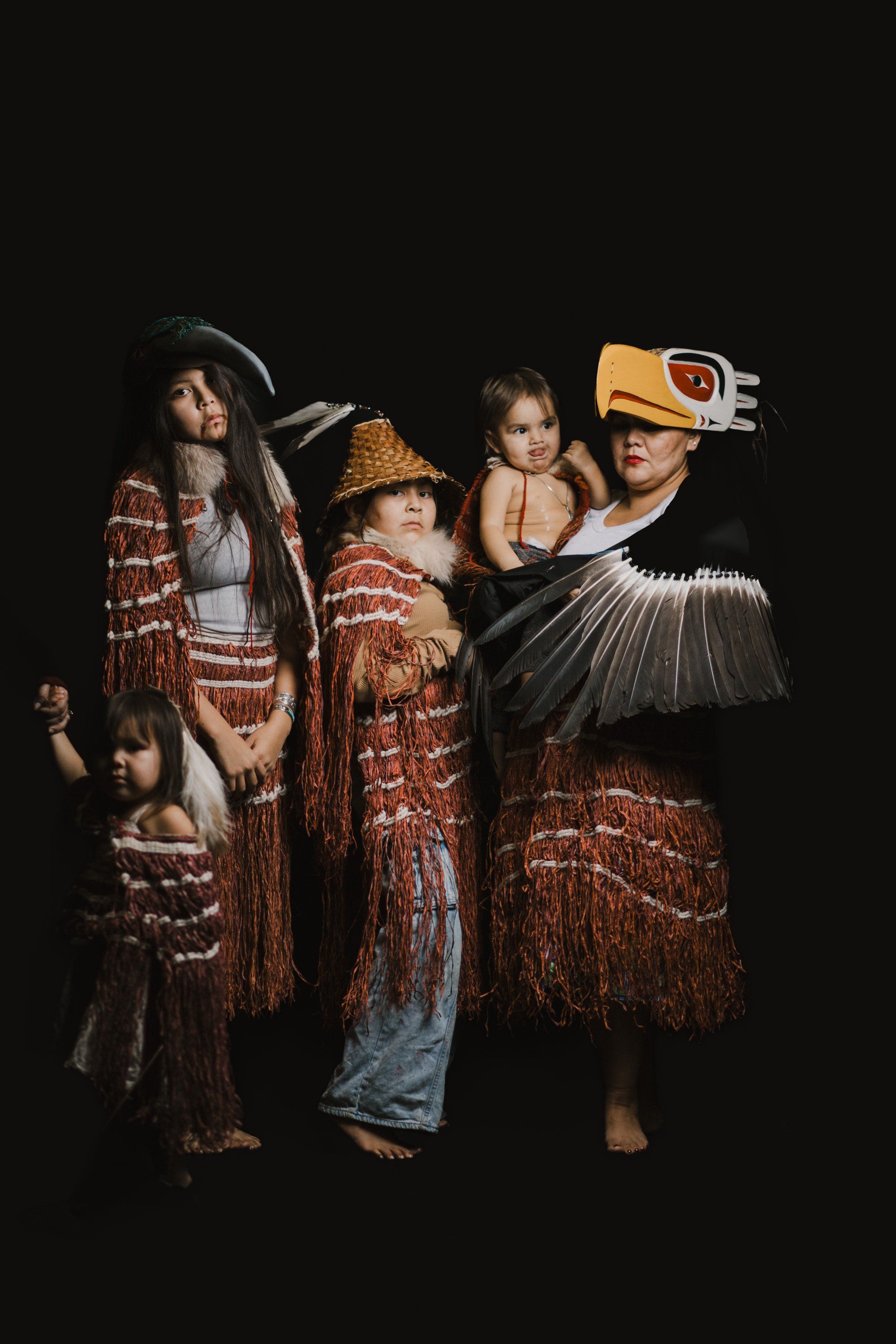 Four Coast Salish family members in traditional regalia with woven blankets and carved masks pose together against a black background