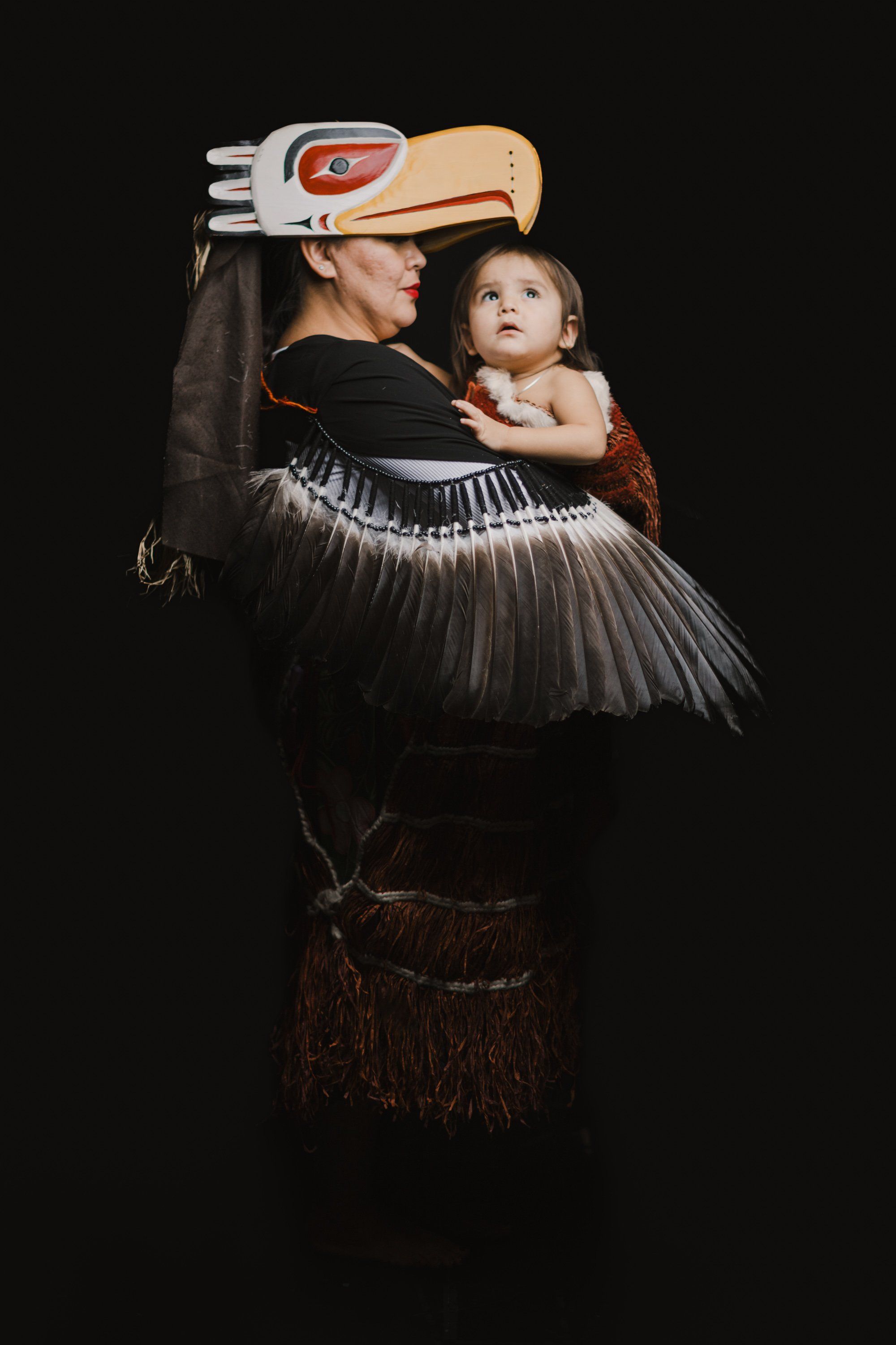 Elder in traditional regalia with carved wooden headdress holds a young child against dark background, their gazes upward in reverent pose