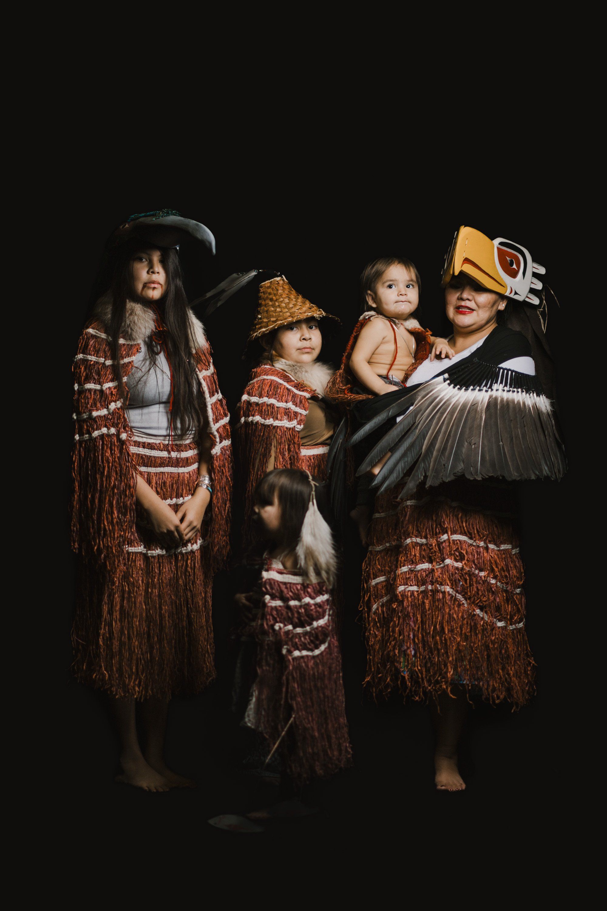 Four people in traditional Coast Salish regalia with fringe and button blankets pose together against a black background, two adults and two children, one ho…