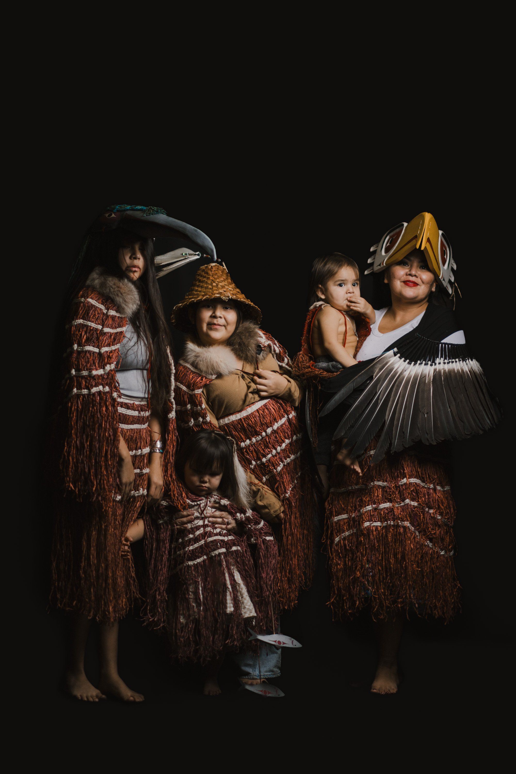 Four Coast Salish family members in traditional regalia with fringed button blankets and carved headdresses pose together against a black background