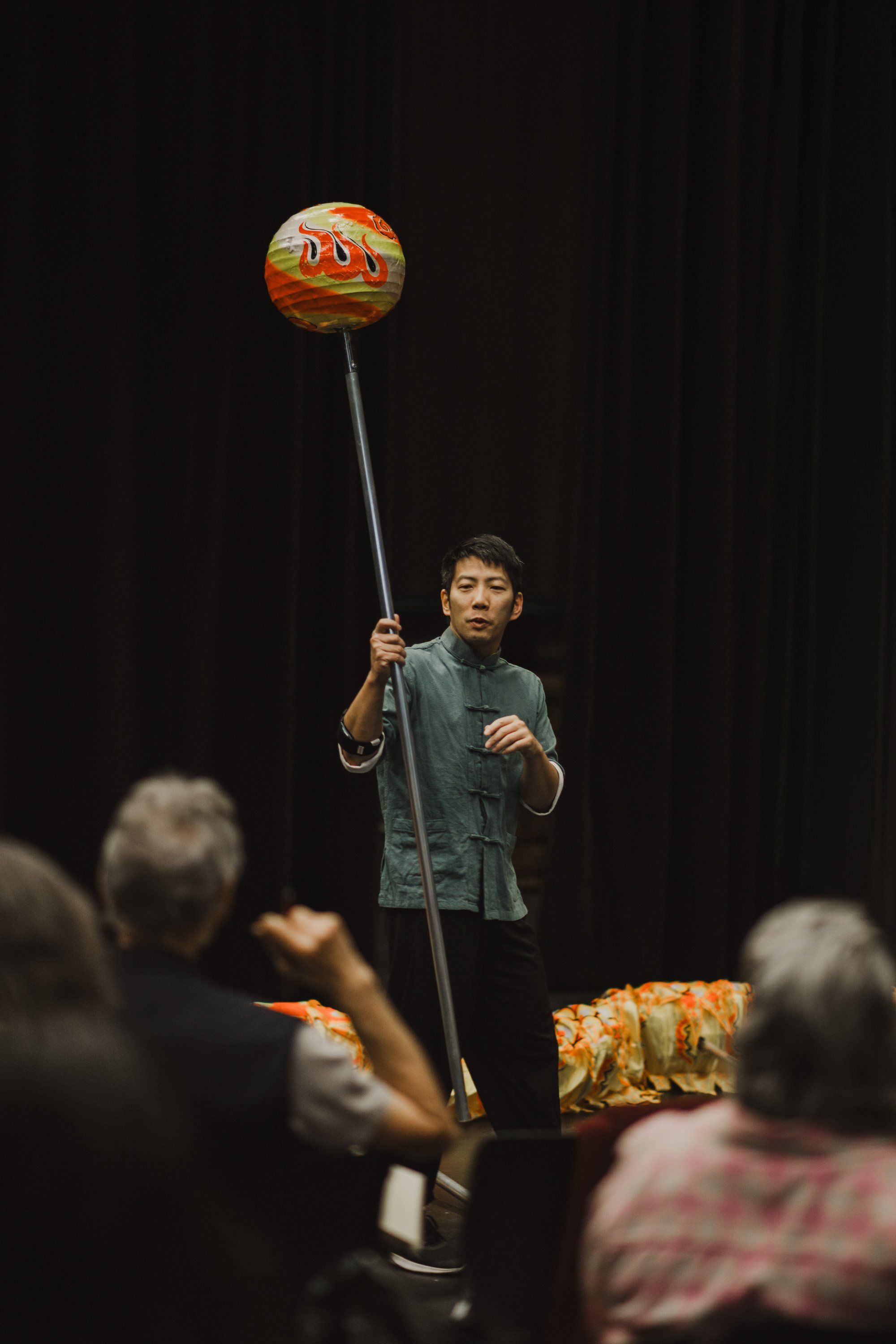 Young performer holds a tall pole with a decorative red and gold button on top while standing on stage before an audience on a dark background