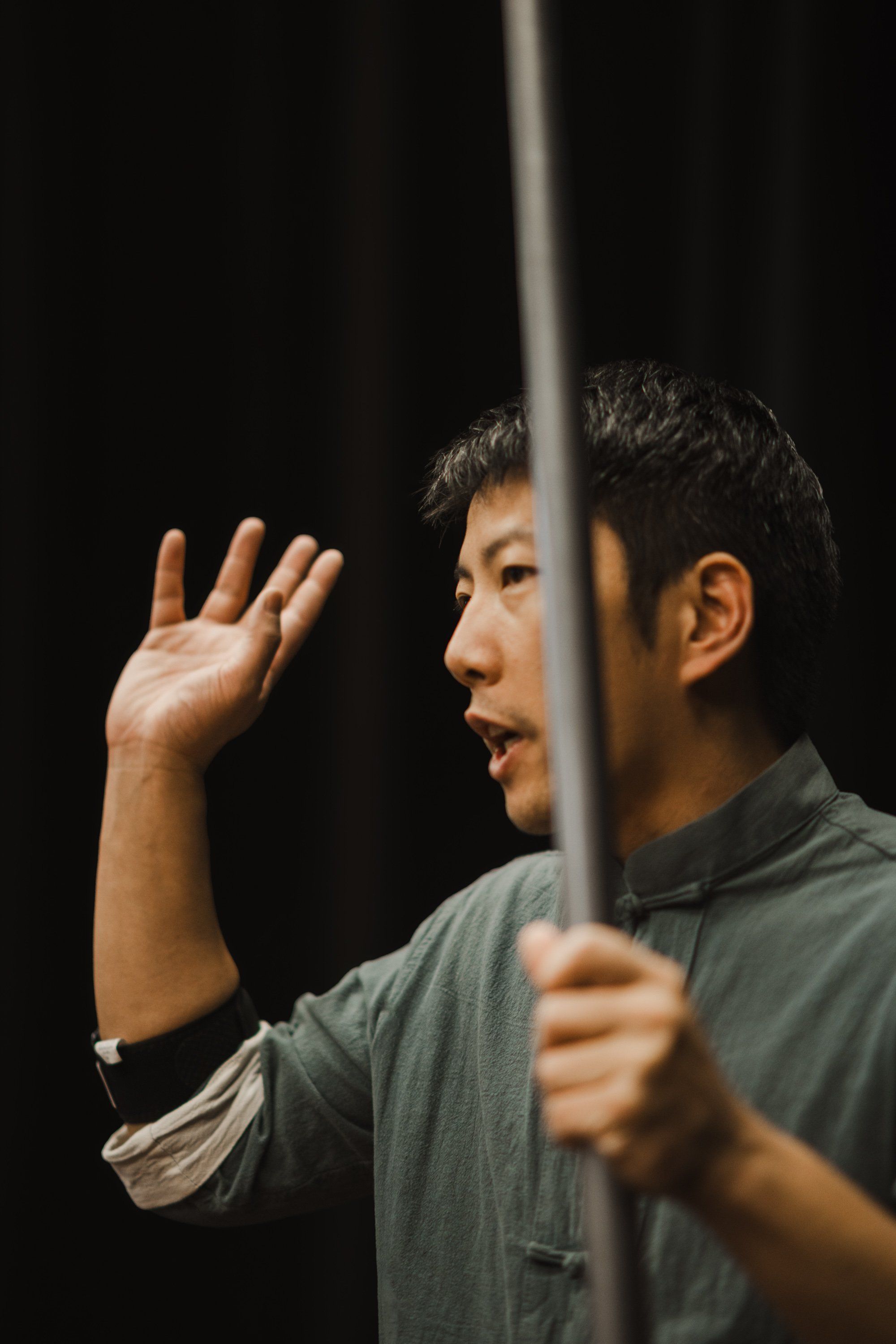 Man in grey shirt gestures expressively while holding a wooden staff, performing against a dark background with dramatic side lighting