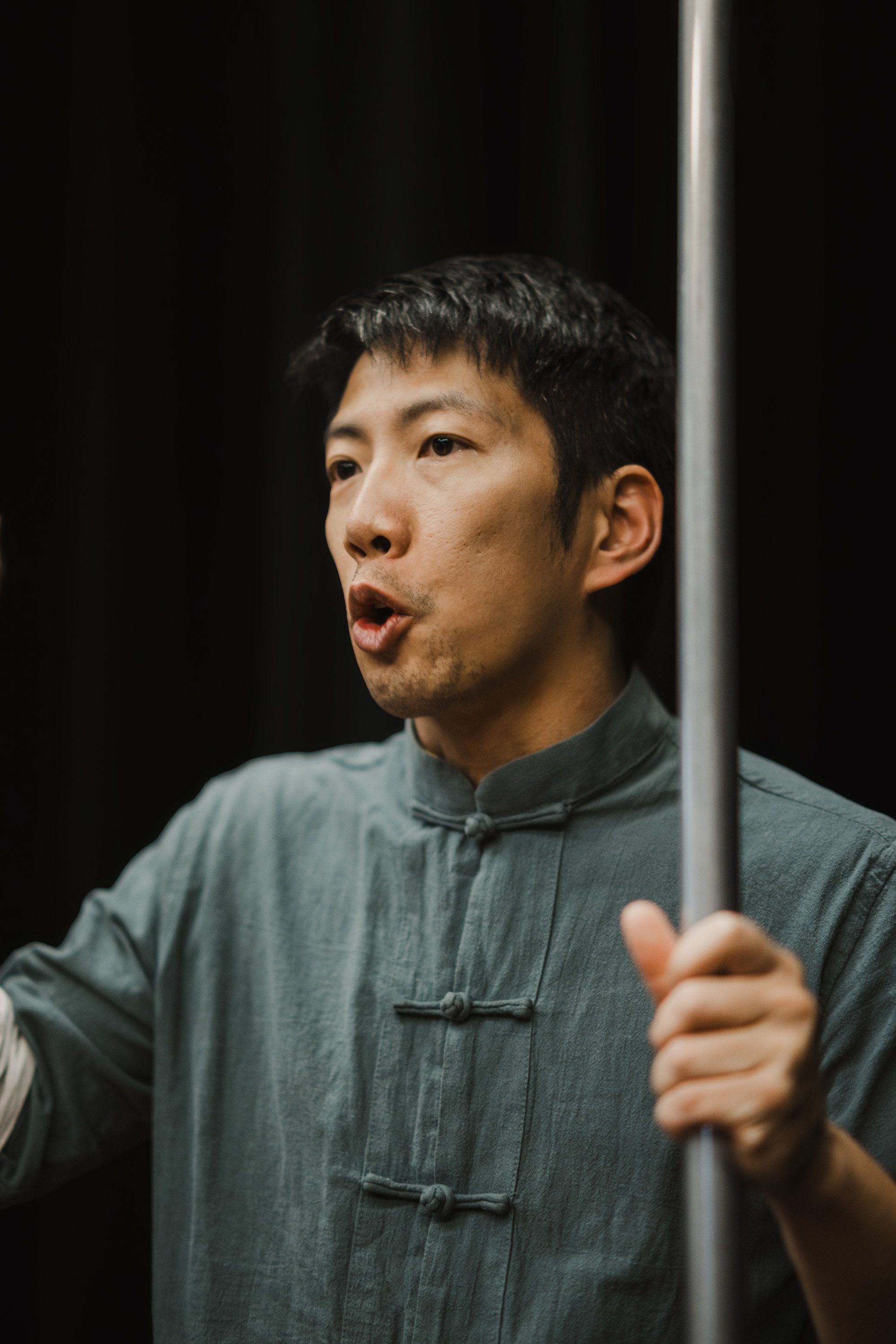 Young man in grey traditional tunic singing with expressive face against dark background, holding wooden staff