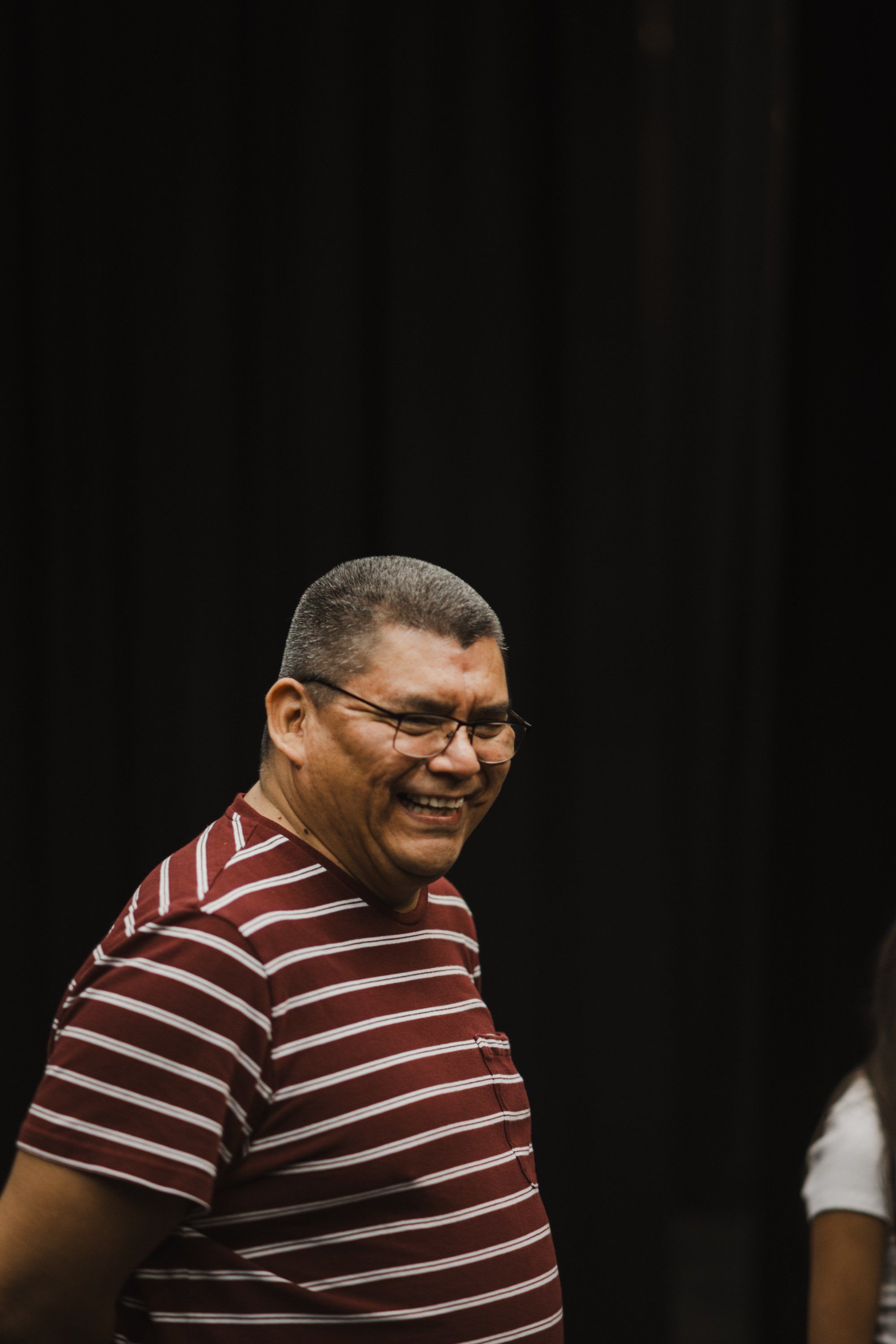 Man in burgundy striped shirt and glasses smiles warmly against a dark backdrop, captured in candid profile during a Coast Salish celebration