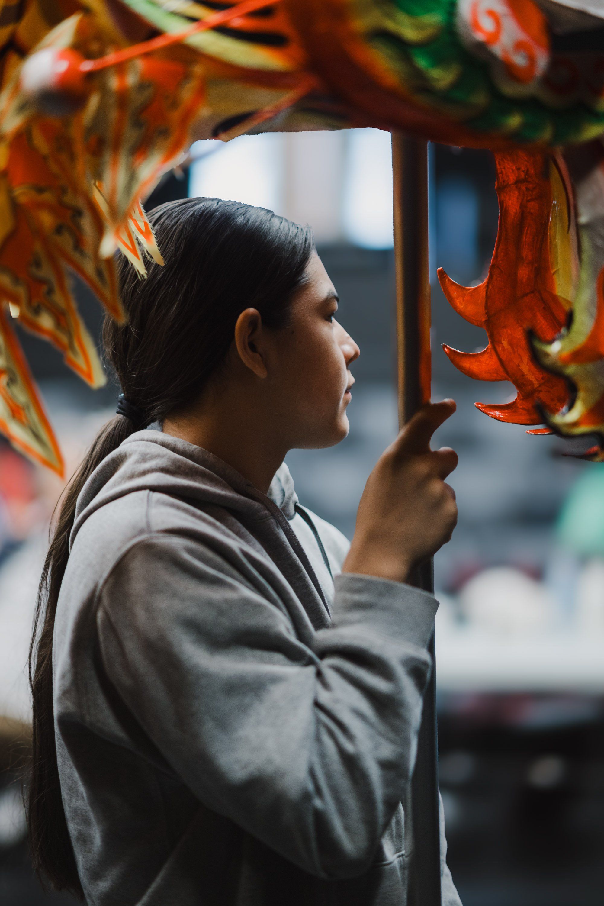 Person in gray hoodie holding wooden staff, profile view, framed by orange and red decorative elements in soft natural light