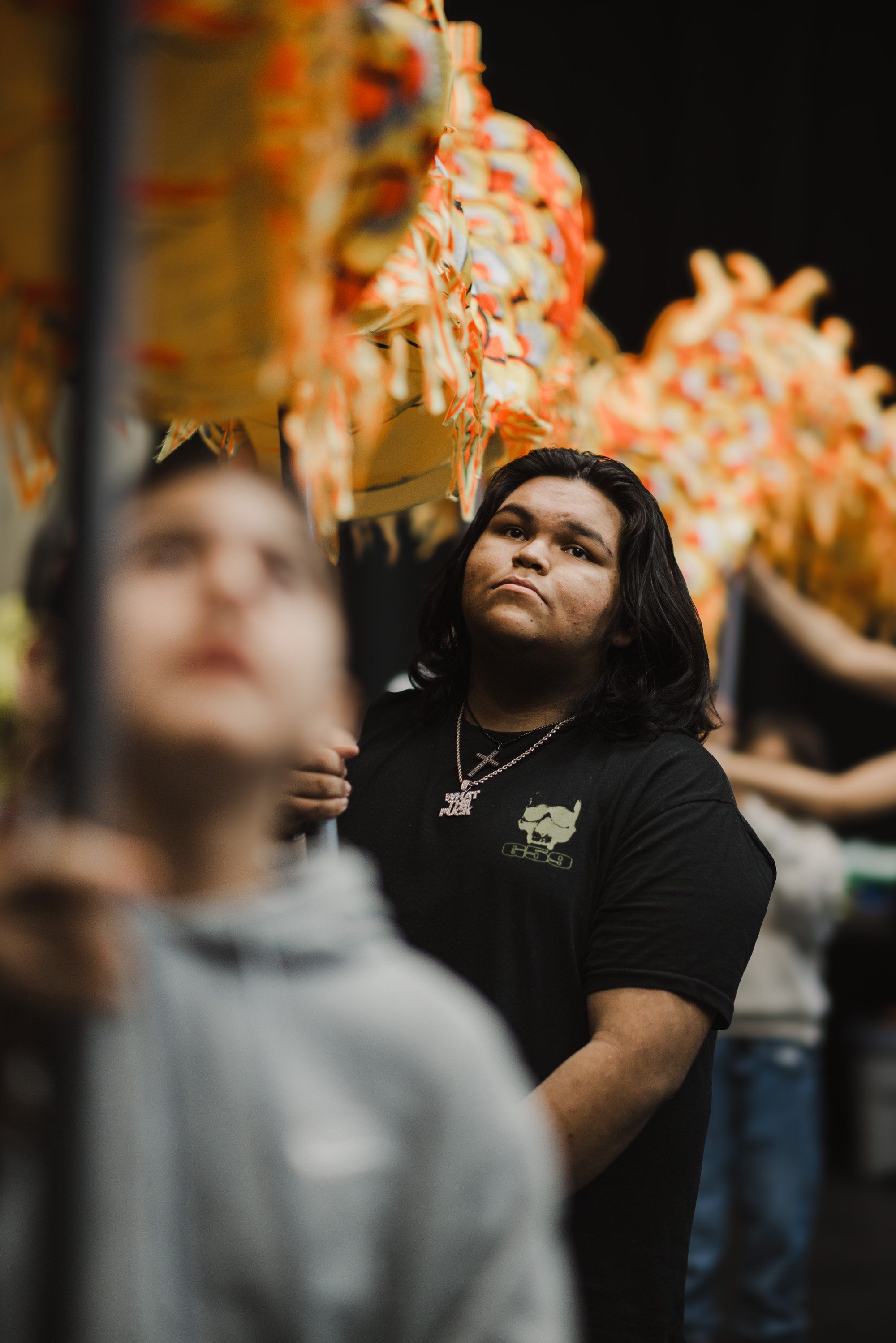 Young man in black shirt gazes upward amid blurred figures and warm amber ceremonial regalia at night