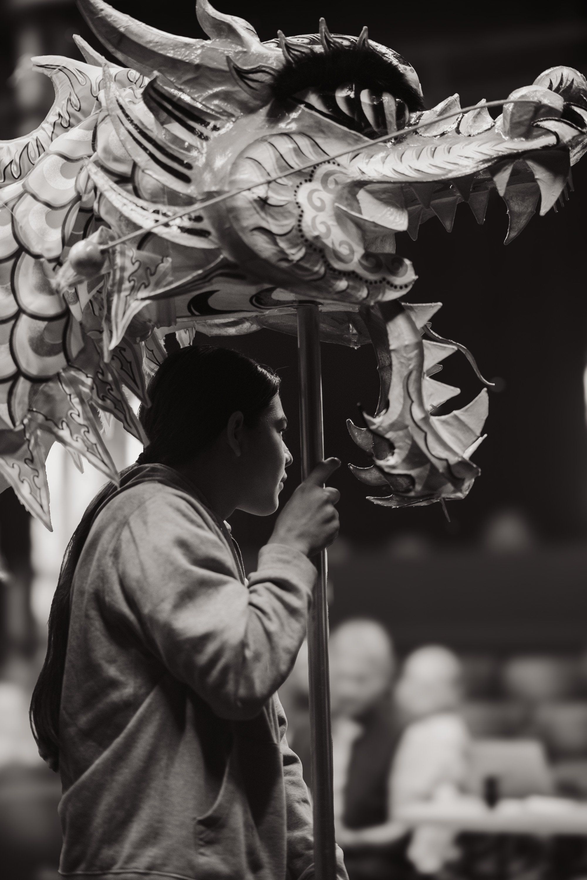 Man in profile holds tall cedar dragon headdress aloft against dark background, intricate carved details illuminated in dramatic black and white