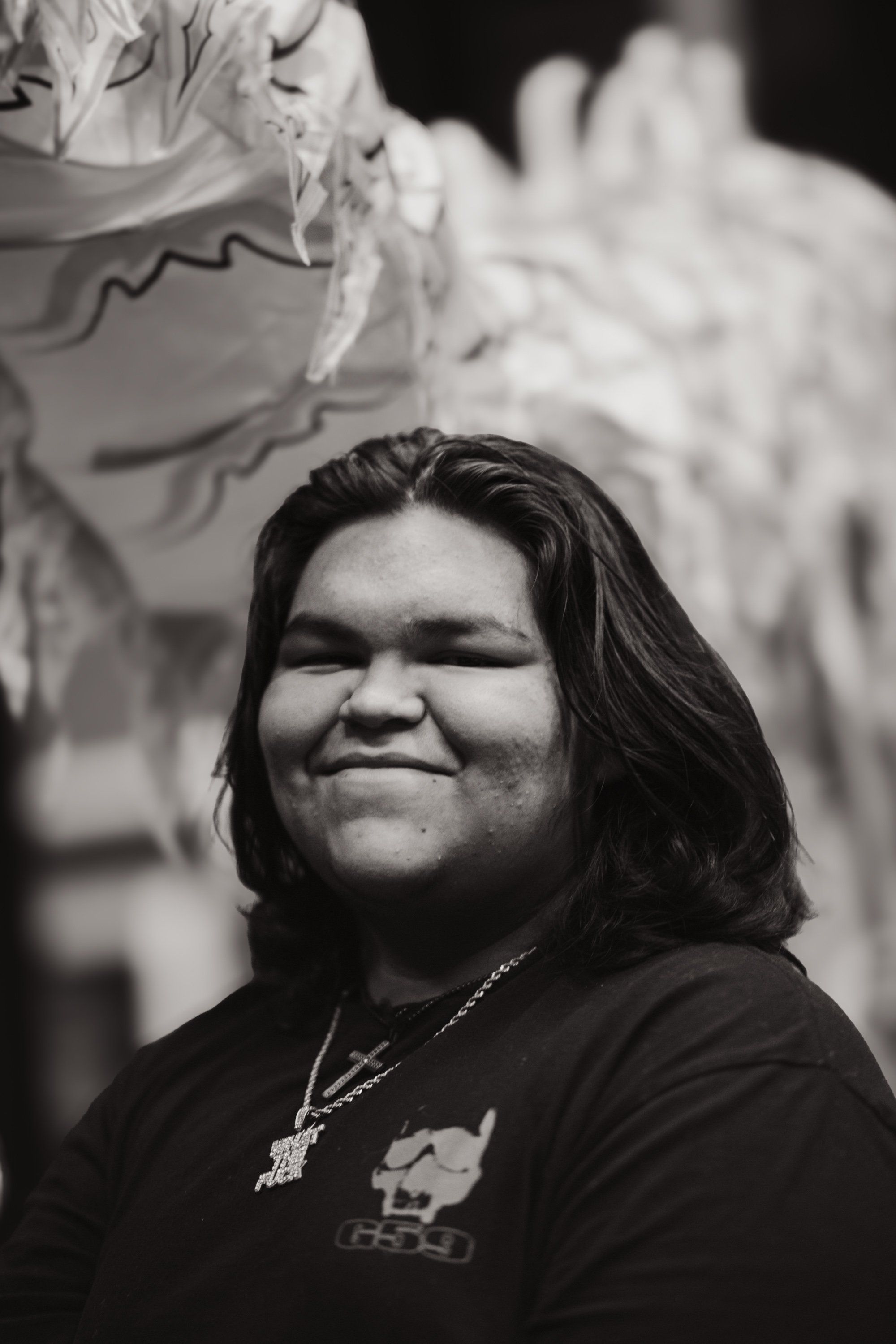 Person with long dark hair wearing a black shirt and pendant, smiling at camera against blurred white ceremonial backdrop in black and white portrait