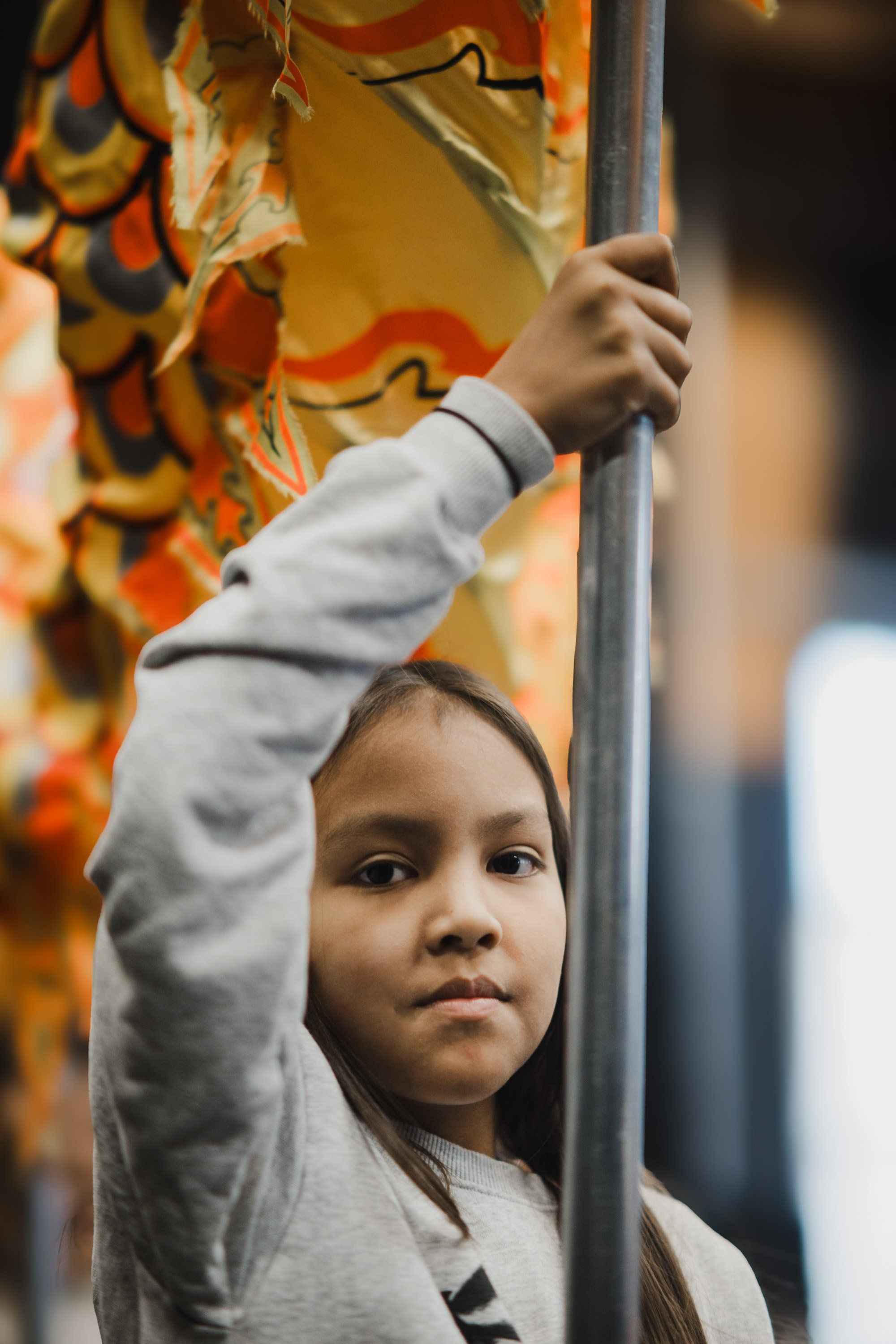 Young child reaching upward on a wooden pole, wearing light gray clothing, with colorful traditional regalia visible in warm golden light behind them