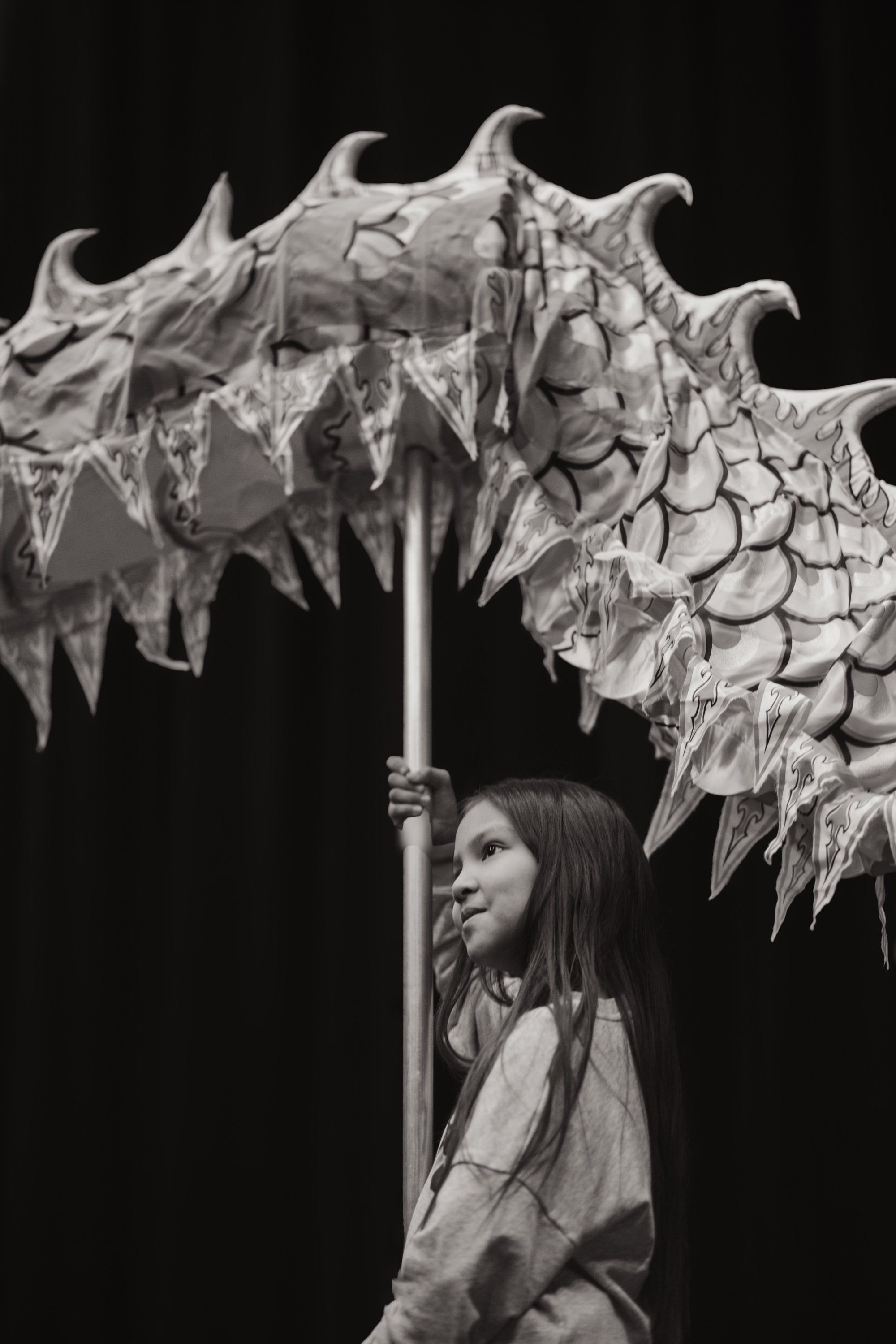 Young dancer holding an ornate carved staff with traditional design against black background, gazing upward in profile