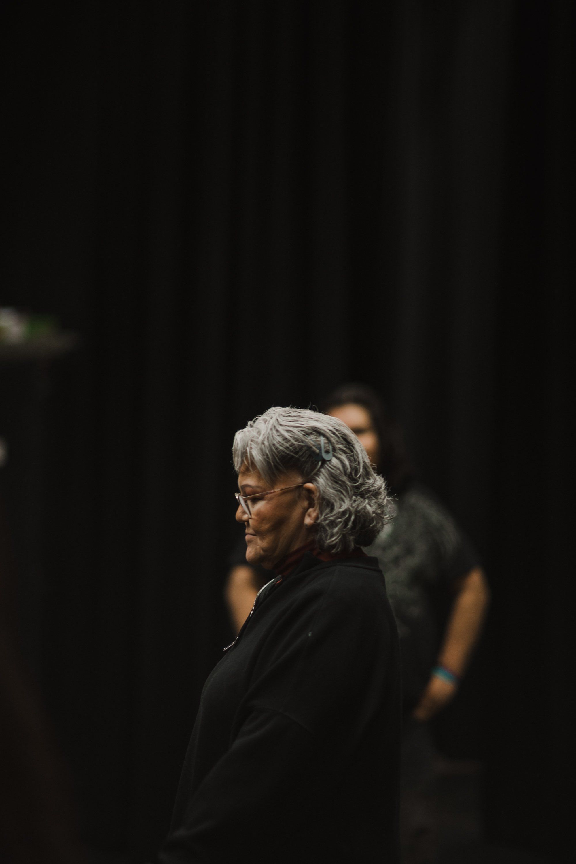 Older woman with gray hair and glasses in profile, wearing black, against dark background with warm stage lighting