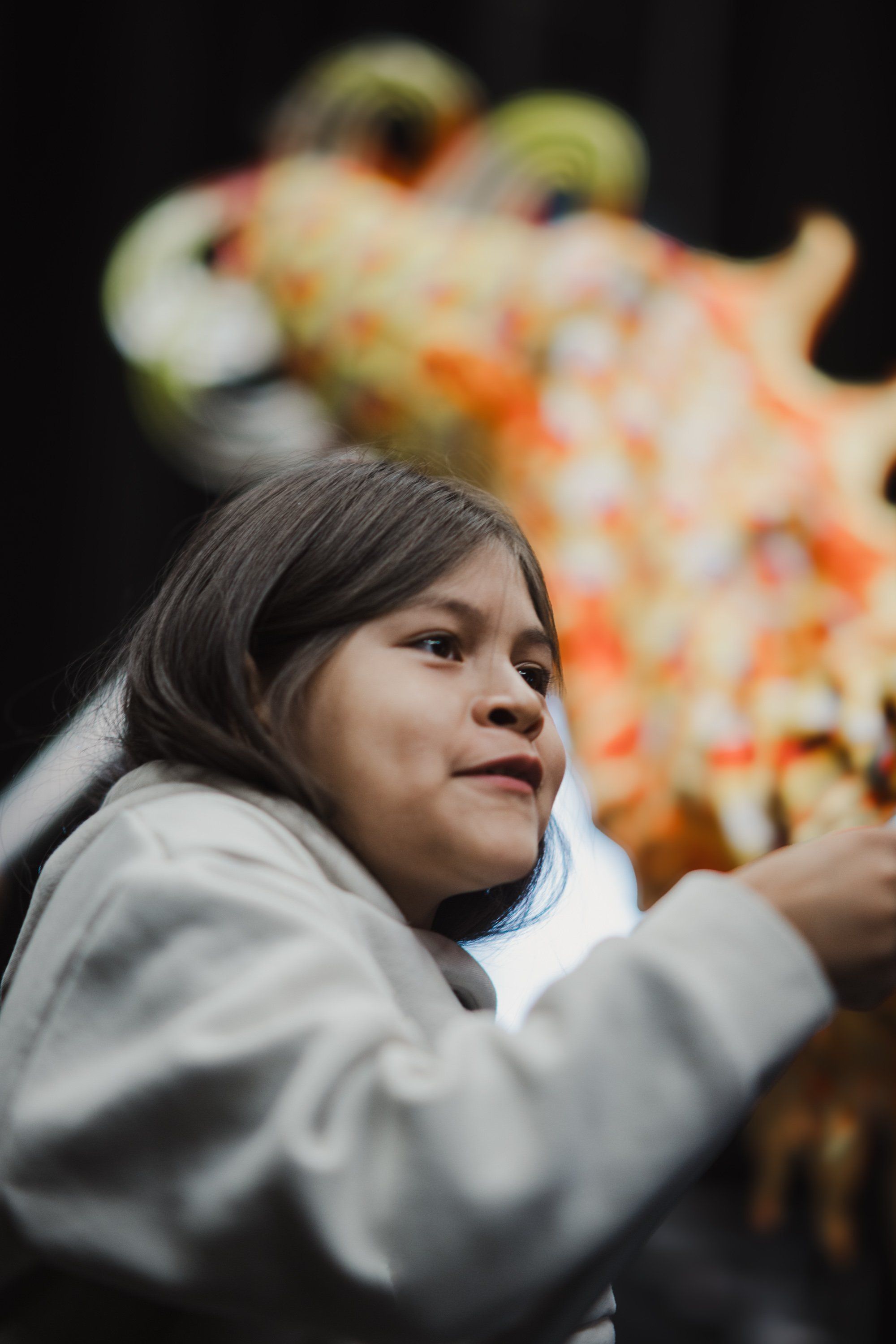 Young girl in white shirt gazes upward with gentle expression, colorful carved figure blurred in background against dark setting