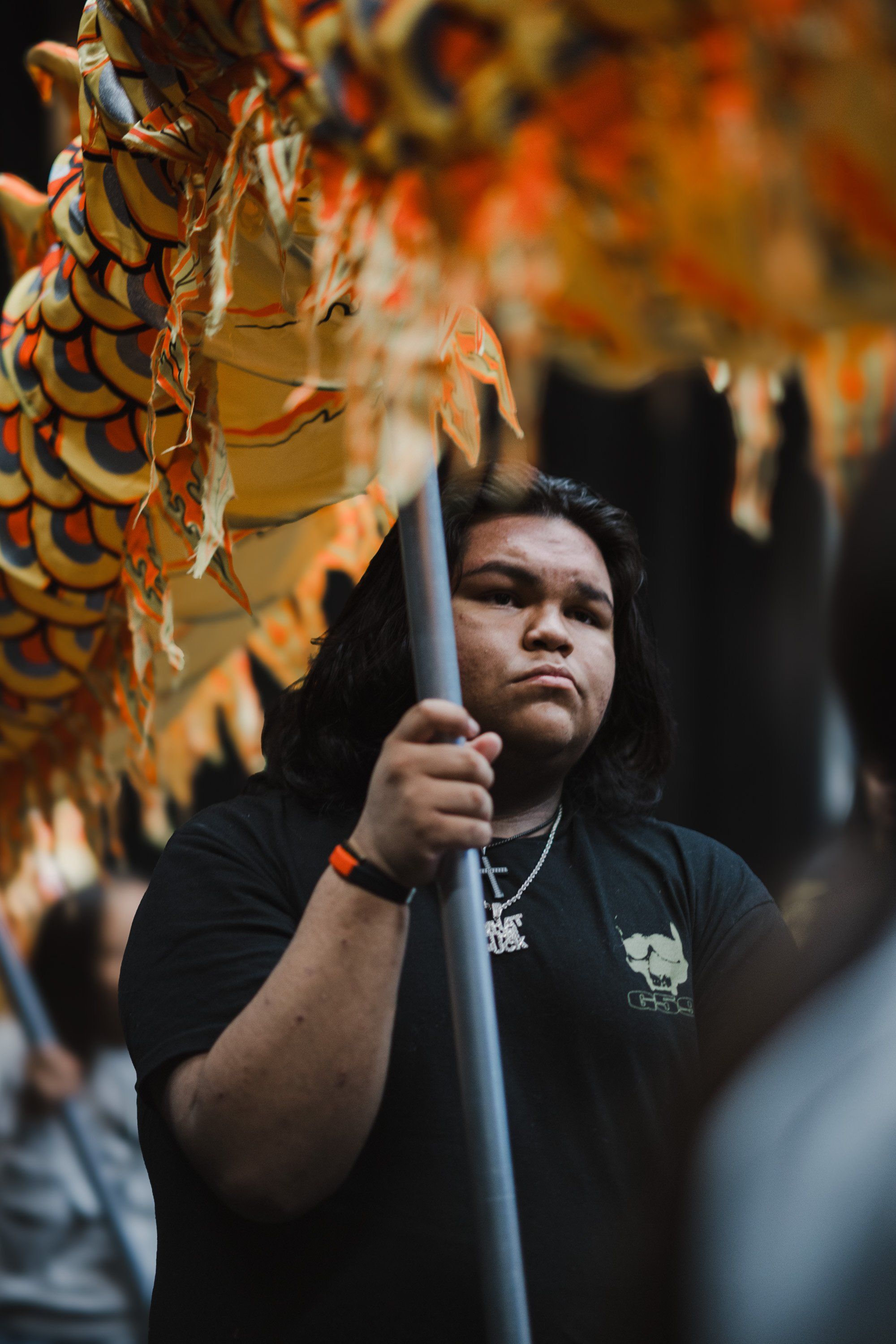 Young man in black shirt holding wooden pole gazes upward at ornate carved wooden mask, warm golden light illuminating the sacred regalia