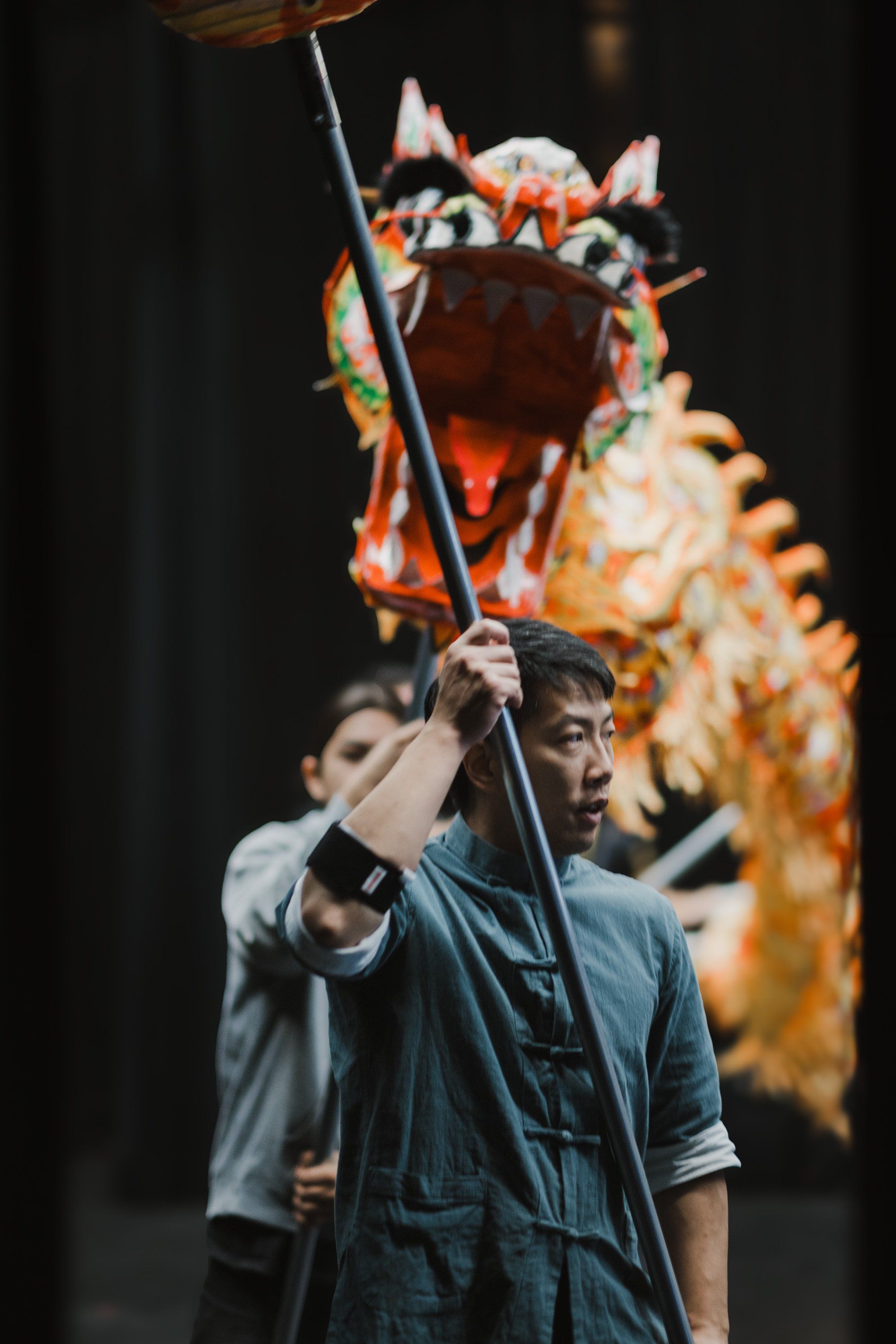 Performer in denim holds a tall pole beneath an ornate wooden wolf mask with orange and white carved details against a dark background