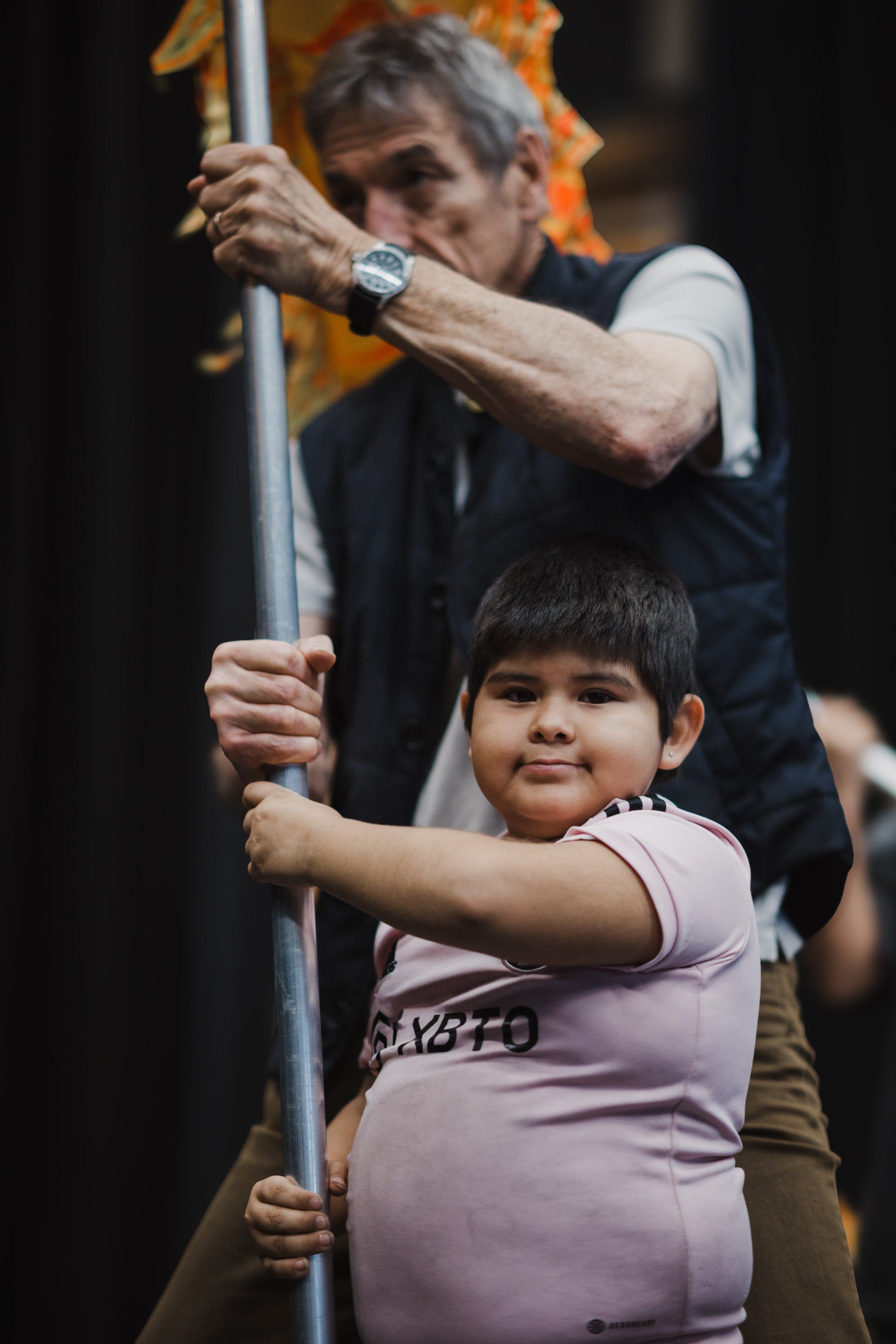 Adult and child hold a wooden pole together during a Coast Salish cultural performance, both smiling warmly at the camera
