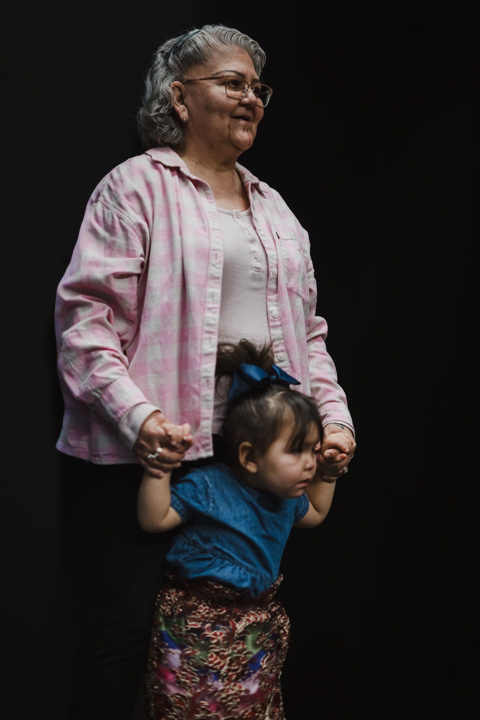An older woman in a pink button-up shirt and a young child in a blue shirt stand together against a black background, holding hands in a moment of quiet conn…