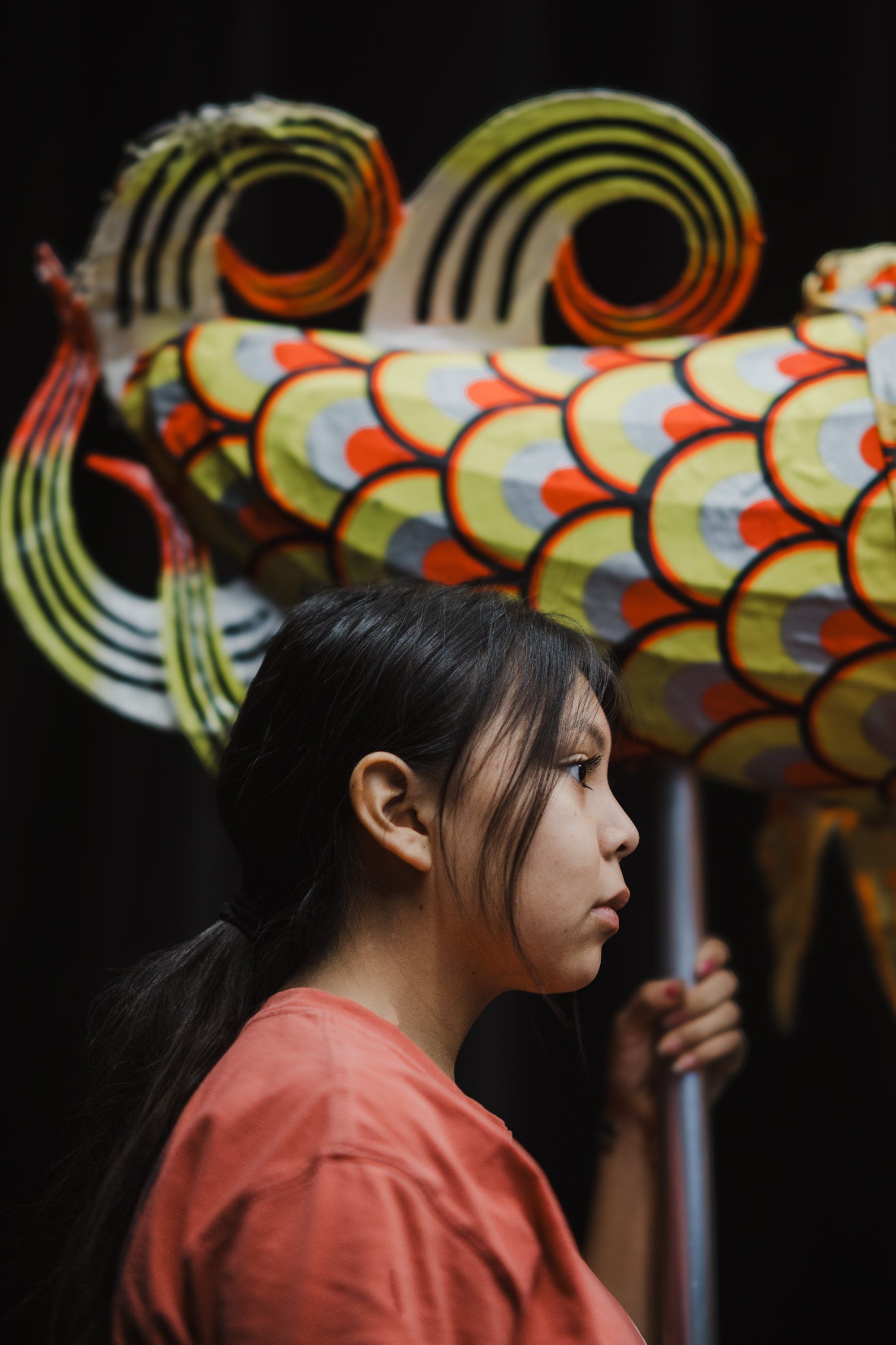 Young woman in coral shirt gazes left, illuminated against a ornate wooden coastal mask with concentric circles and vibrant red-orange details