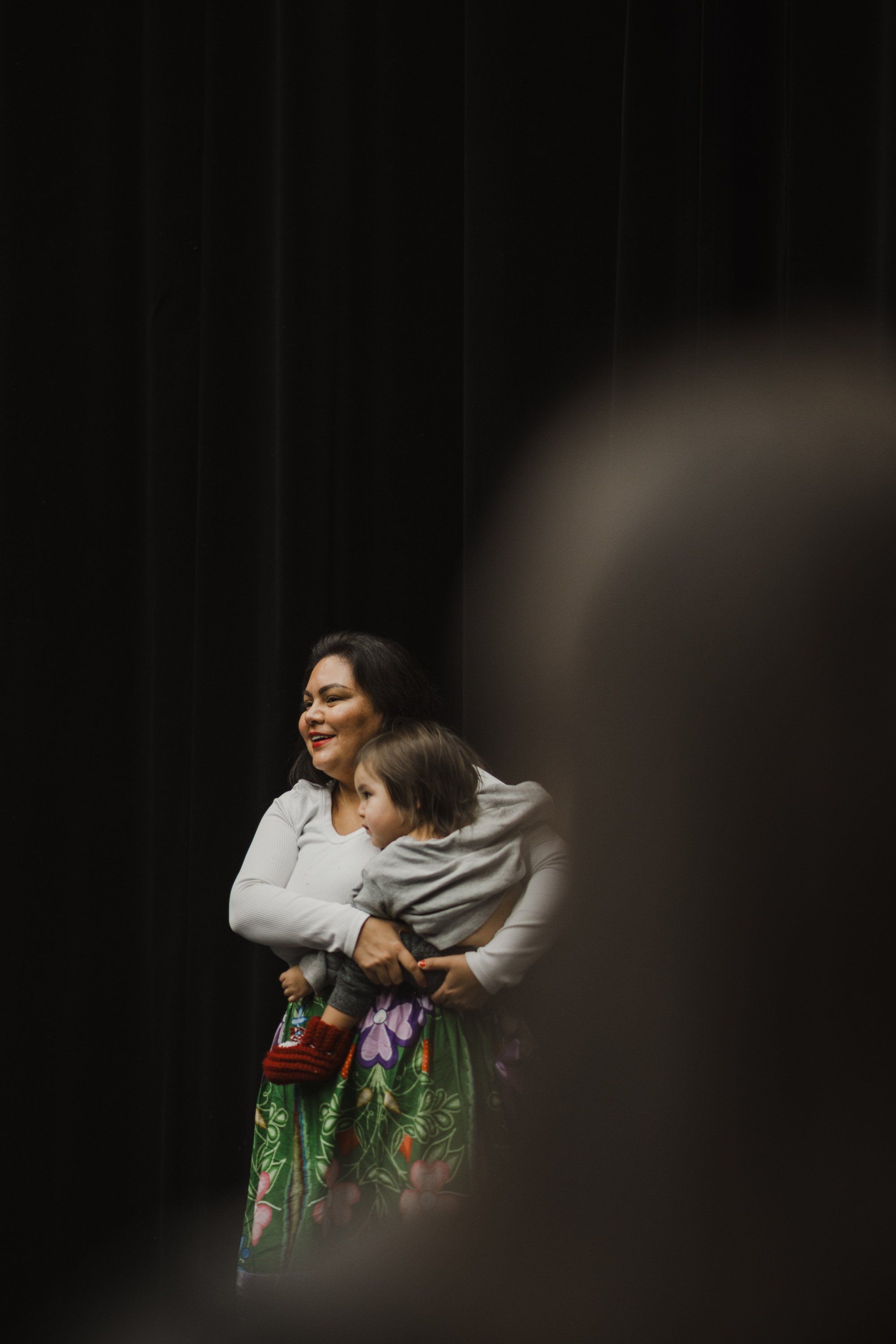 Woman in white shirt holding child, both smiling, against dark background with warm theatrical lighting