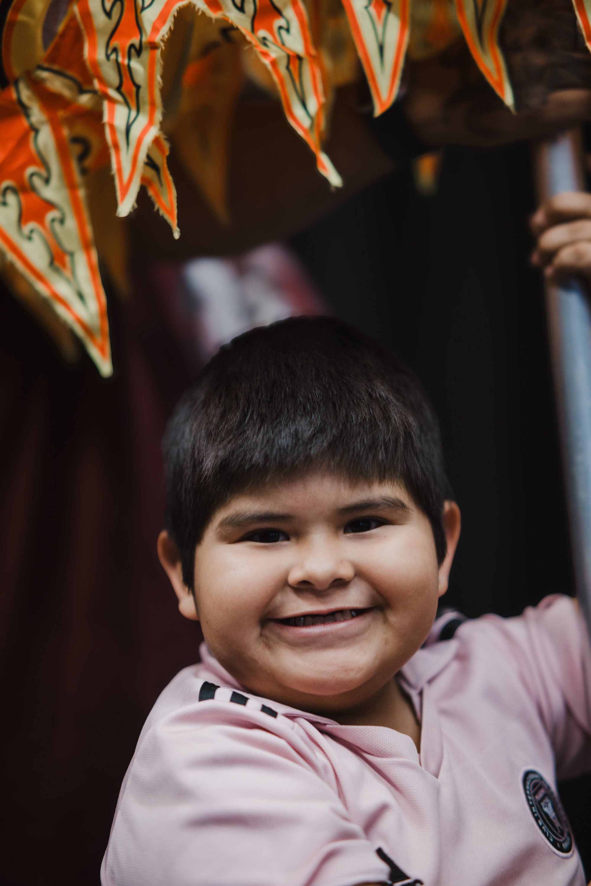 Young child in pink shirt smiling beneath ornate orange and teal ceremonial regalia at a Coast Salish cultural celebration