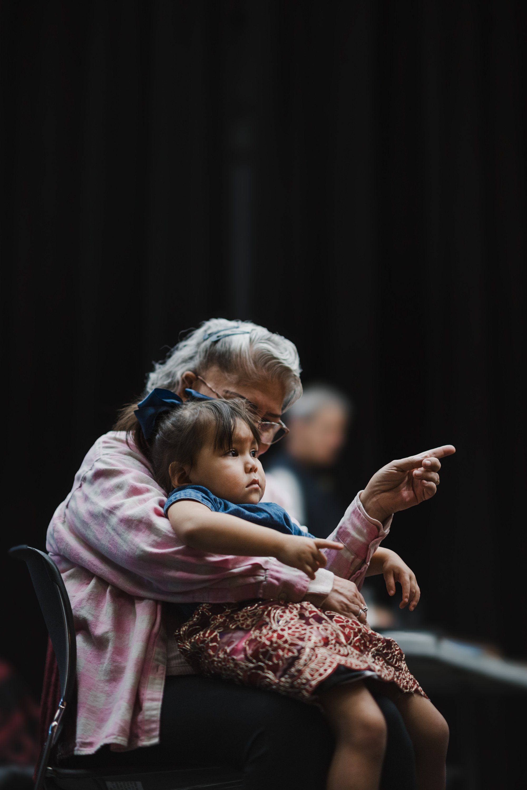Elder and young child seated together pointing forward, dark background, intimate moment during Coast Salish performance